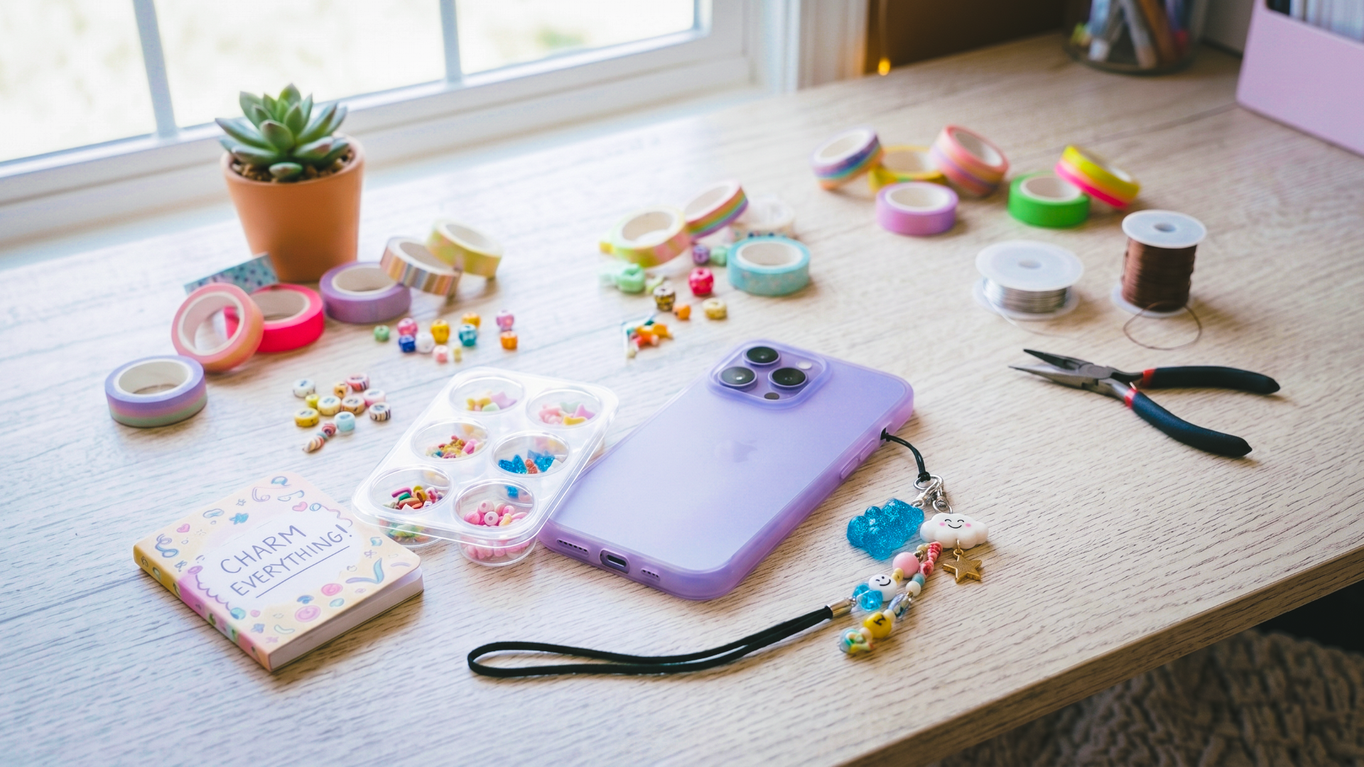 Light wood desk showing a smartphone in a purple case with a charm keychain attached. The table also has washi tape, beads, wire, and needle nose pliars, all set up for a charm bar party for tweens and teens..