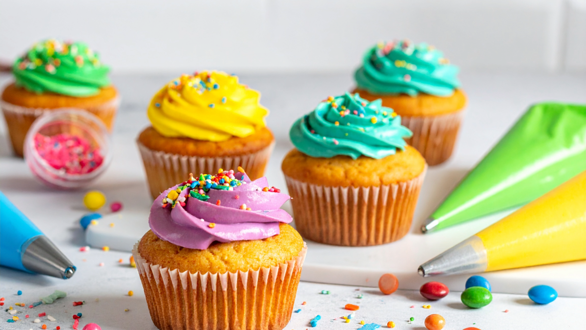 Five vanilla cupcakes with bright swirls of purple, yellow, teal, and green frosting and rainbow sprinkles on a white surface with piping bags and colorful candies.