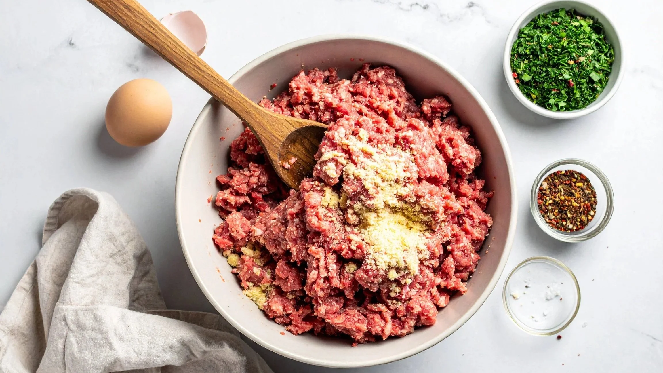 Raw meatball ingredients for game day meatball appetizers ready to be mixed. There is a bowl of ground beef with breadcrumbs and egg already inside. Small bowls of red pepper and garnish, along with a wooden spoon, an egg, and a dish towel.