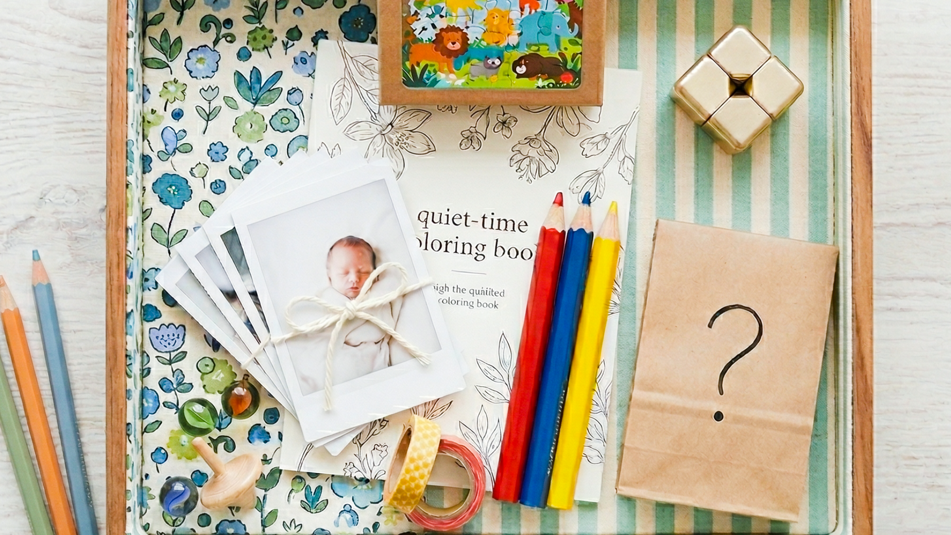 A curated DIY boredom box for kids featuring a small puzzle, baby photos, a coloring book, and a mystery surprise bag in a fabric-lined wooden tray.