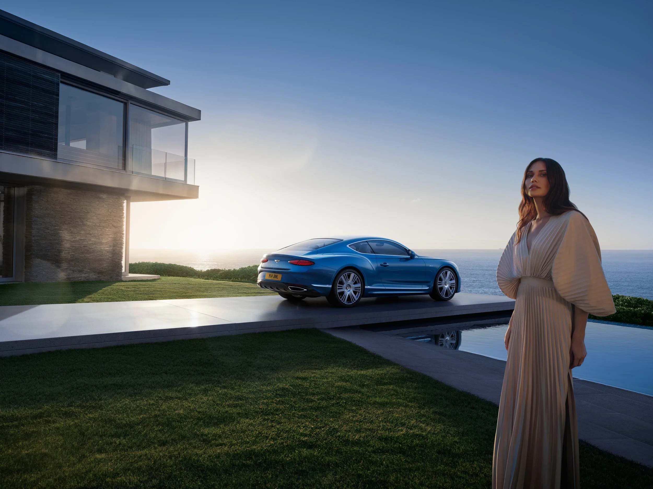 Woman standing outside near a modern house with a blue sports car parked on a driveway, overlooking the water during sunset.
