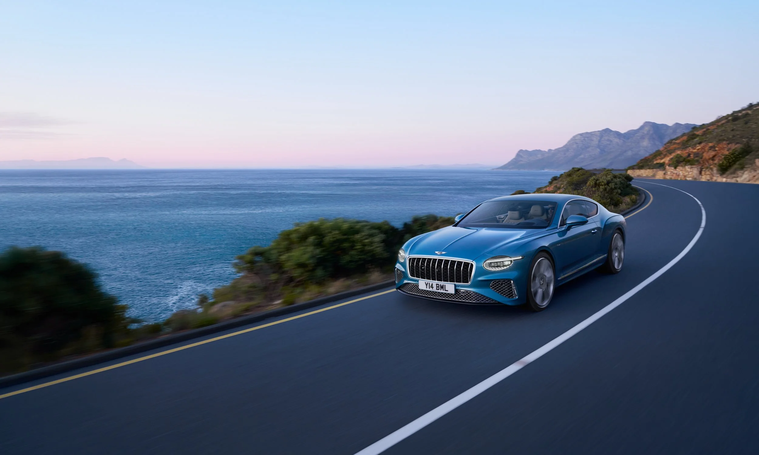 A blue luxury sports car driving along a coastal road with ocean on one side and mountains on the other at sunset.