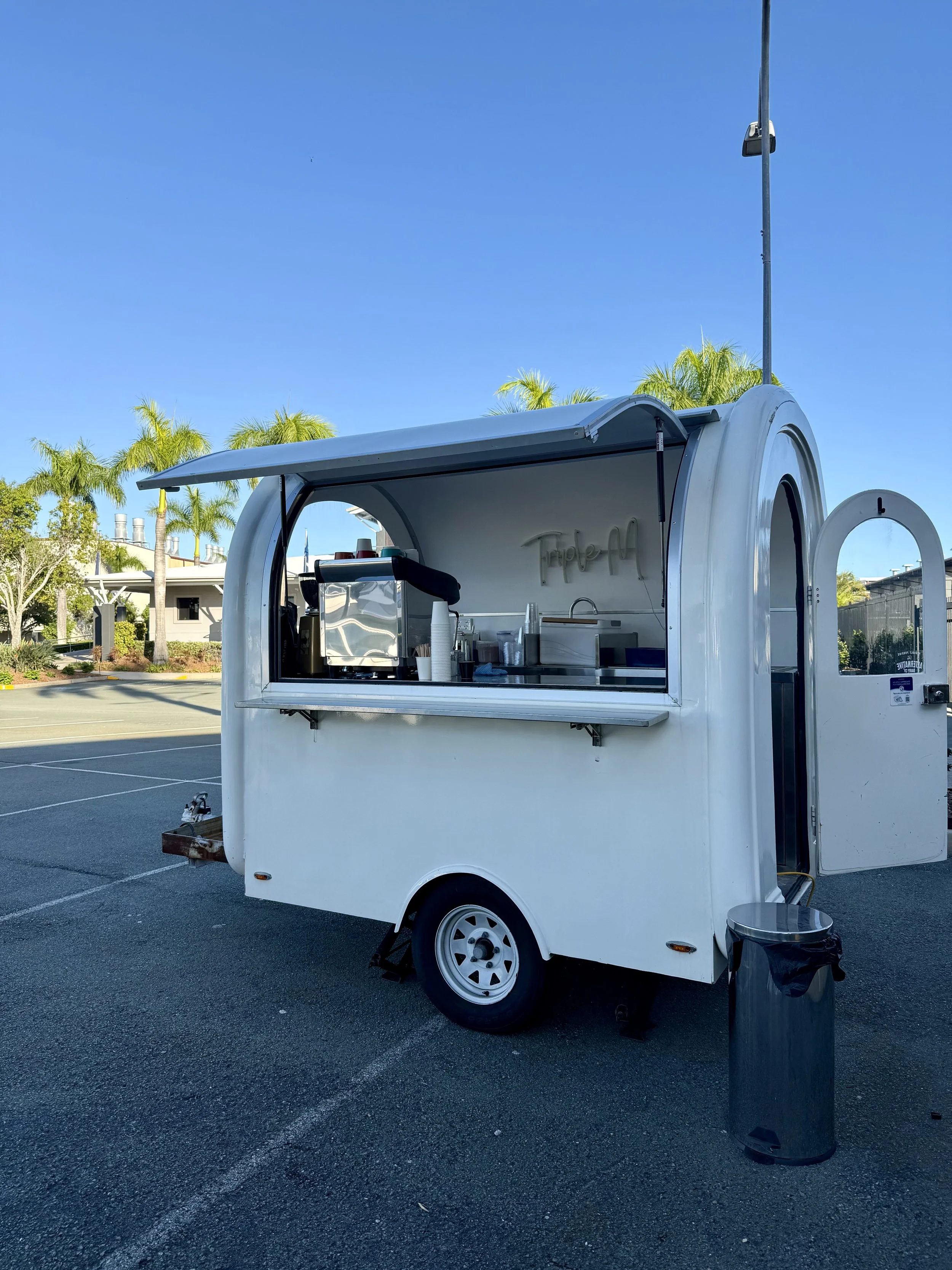 A white mobile food trailer with an open serving window, parked in a lot with palm trees in the background and a clear blue sky overhead.
