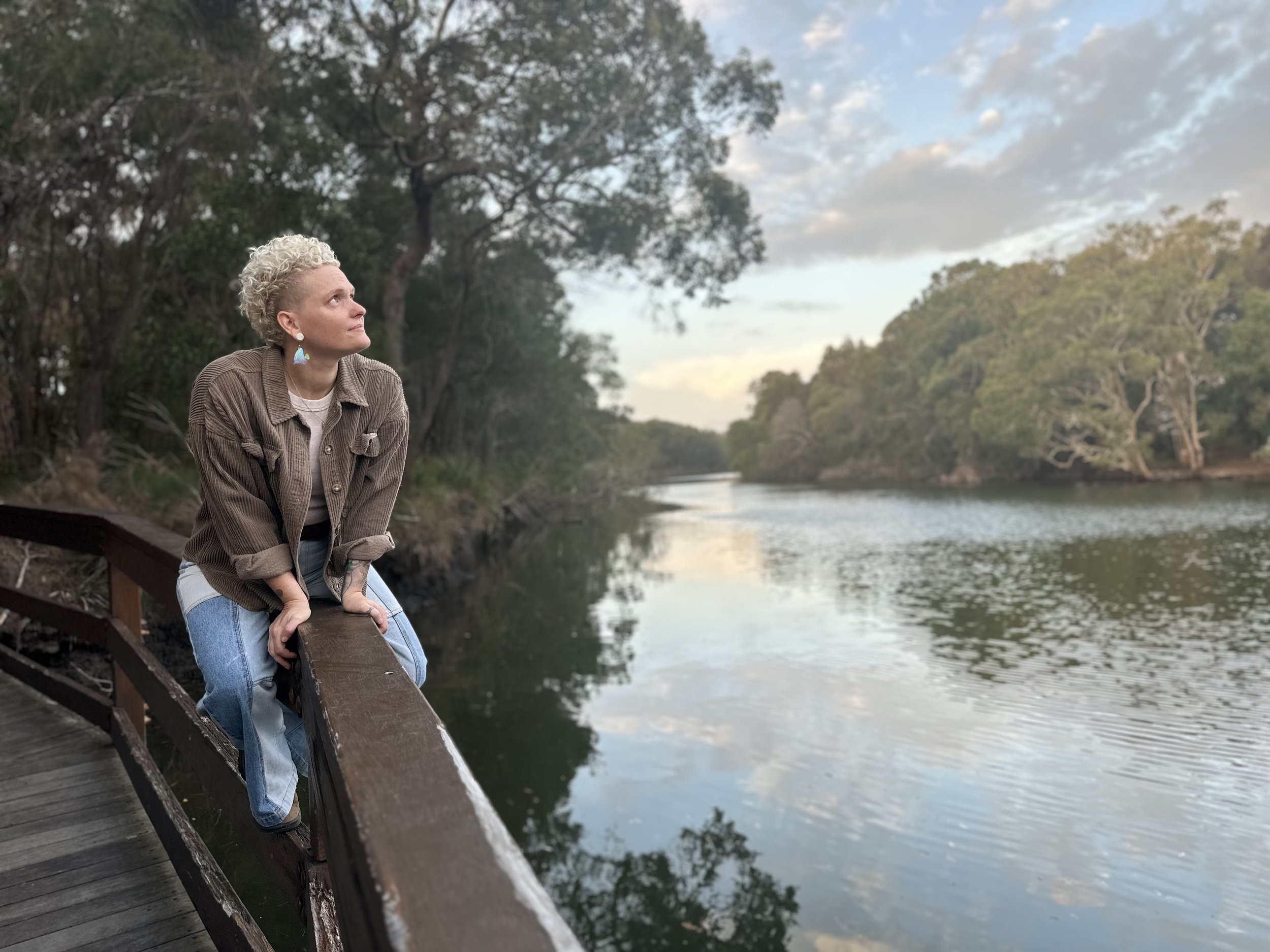 A woman with short curly blond hair and earrings sitting on a wooden bridge by a river, surrounded by trees and a partly cloudy sky.