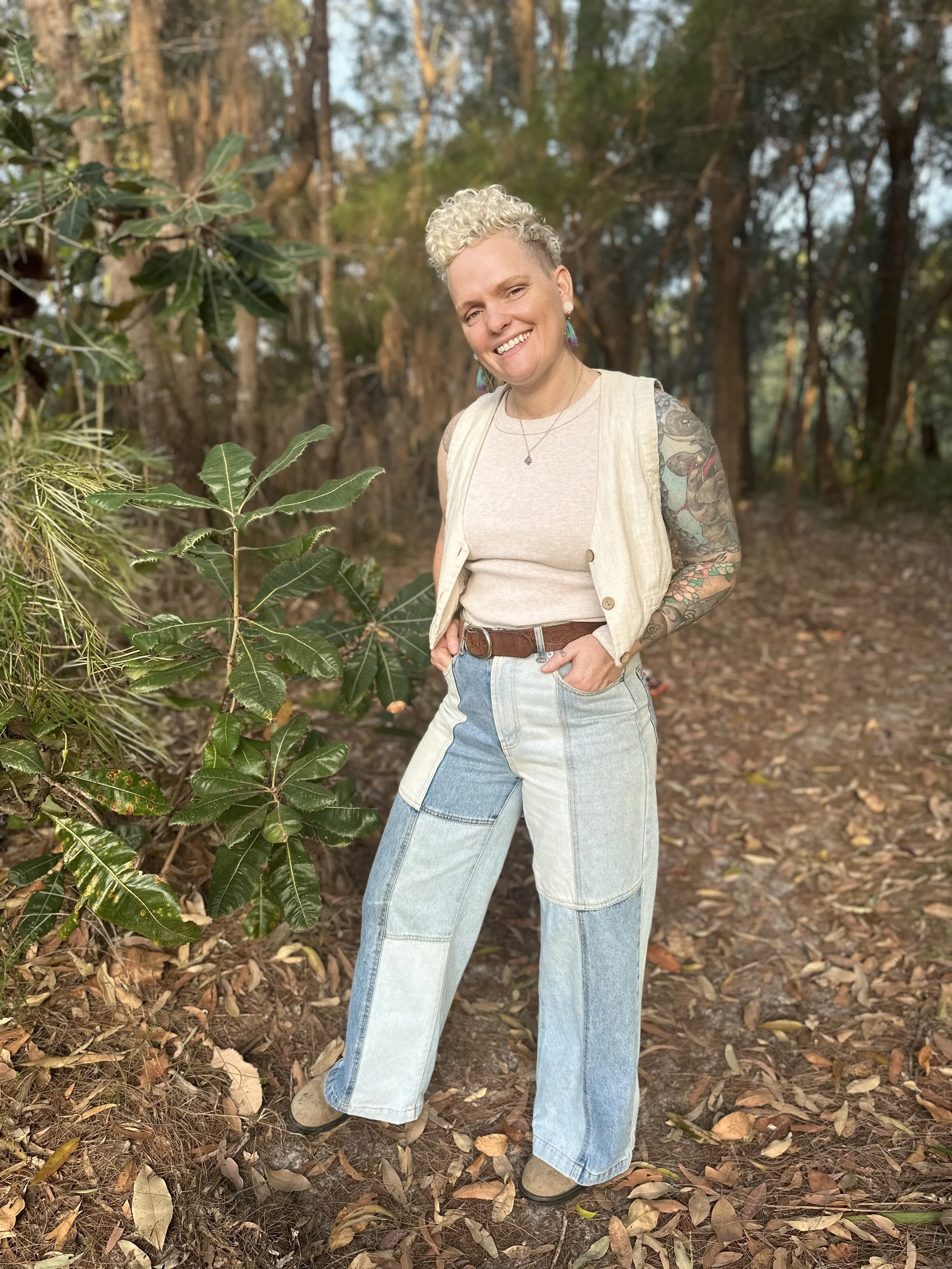 A person with curly blonde hair and tattoos on their arms standing outdoors in a wooded area, smiling and wearing a beige sleeveless vest over a light shirt and patchwork jeans.