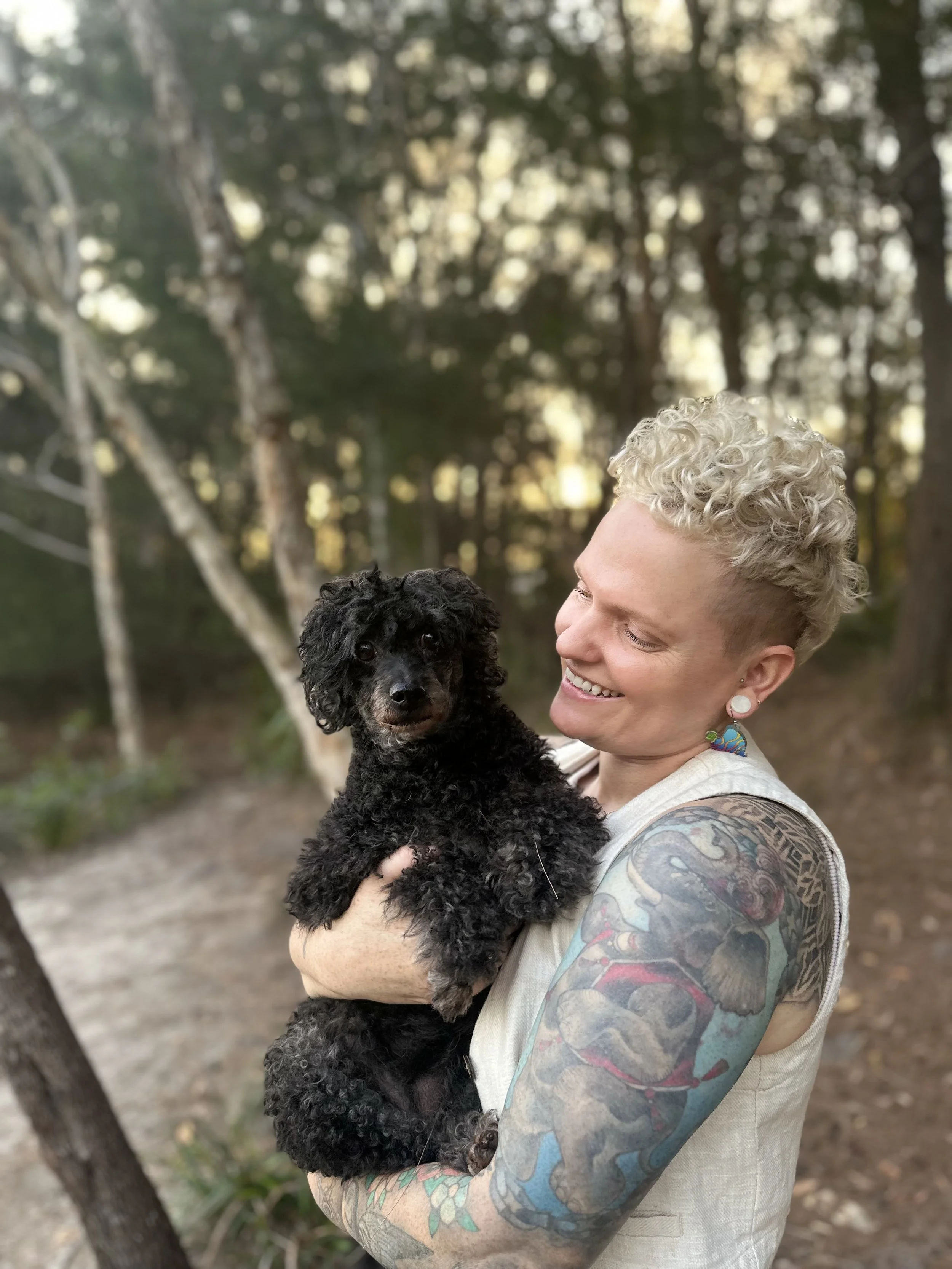 A woman with short, curly blonde hair smiling and holding a black curly-haired puppy outdoors in a forested area.