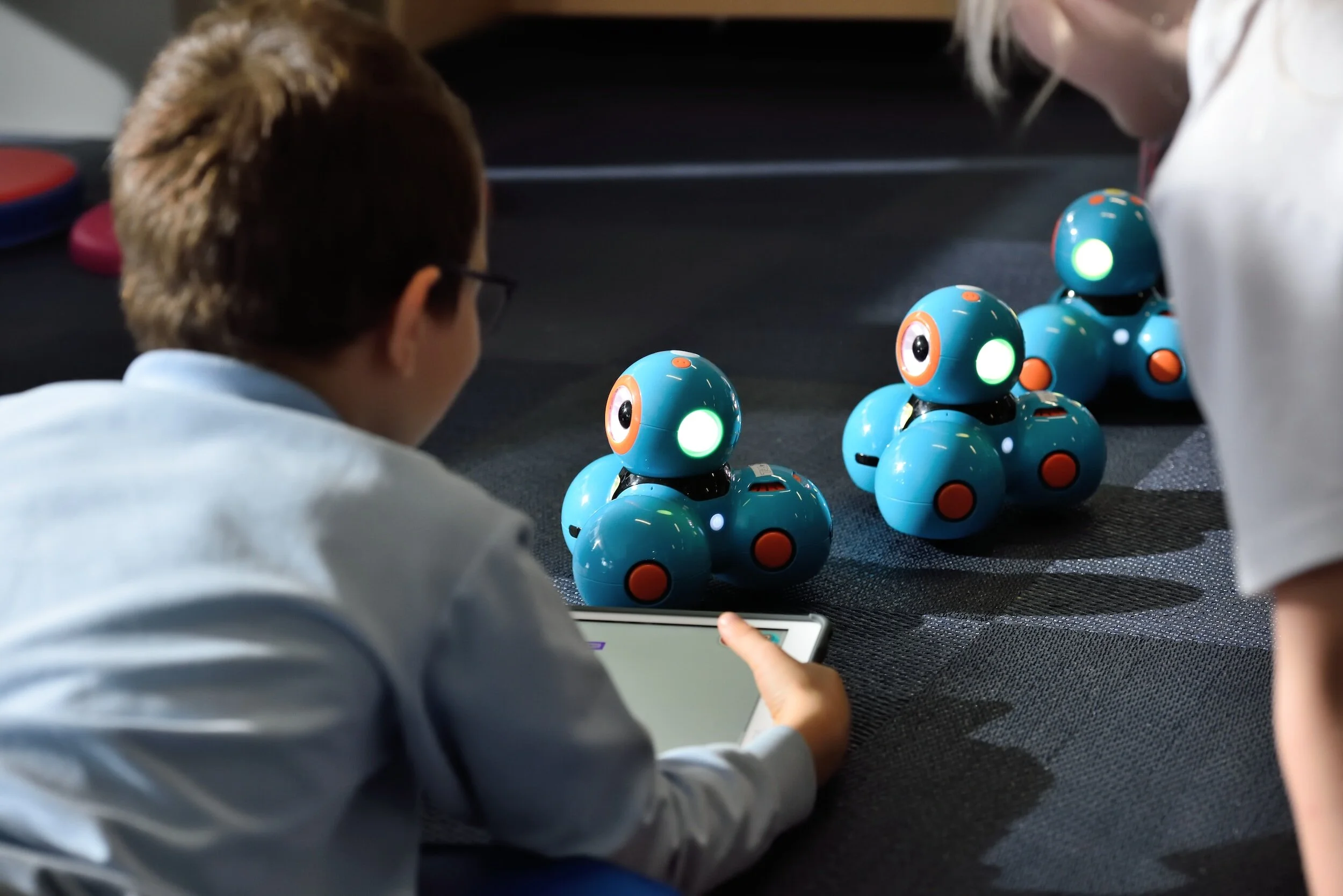 Children interacting with small blue robots using a tablet on a carpeted floor.