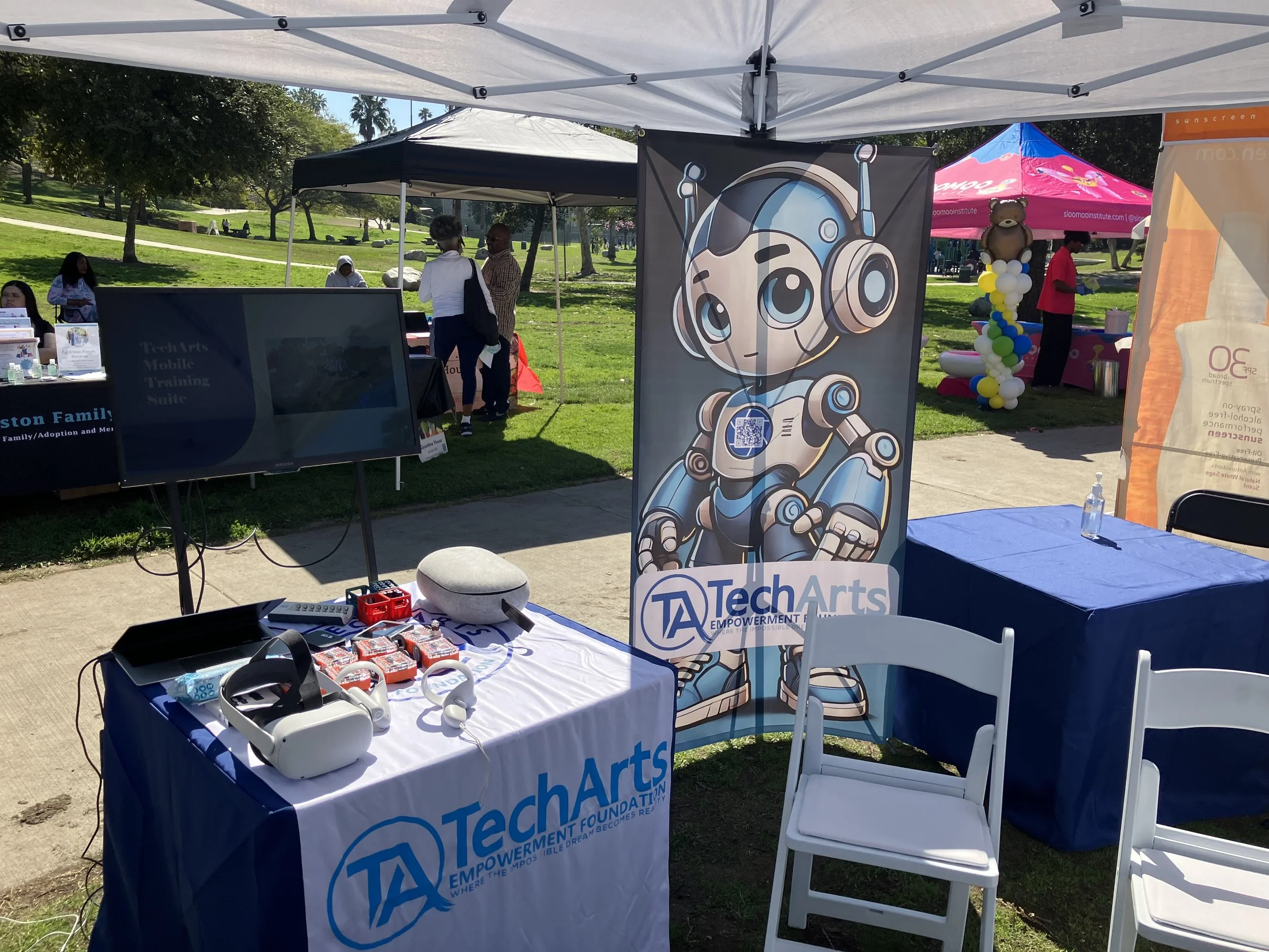 Tech Arts booth at outdoor event with banners, VR headset, pamphlets, and promotional items under a canopy tent in a park setting.