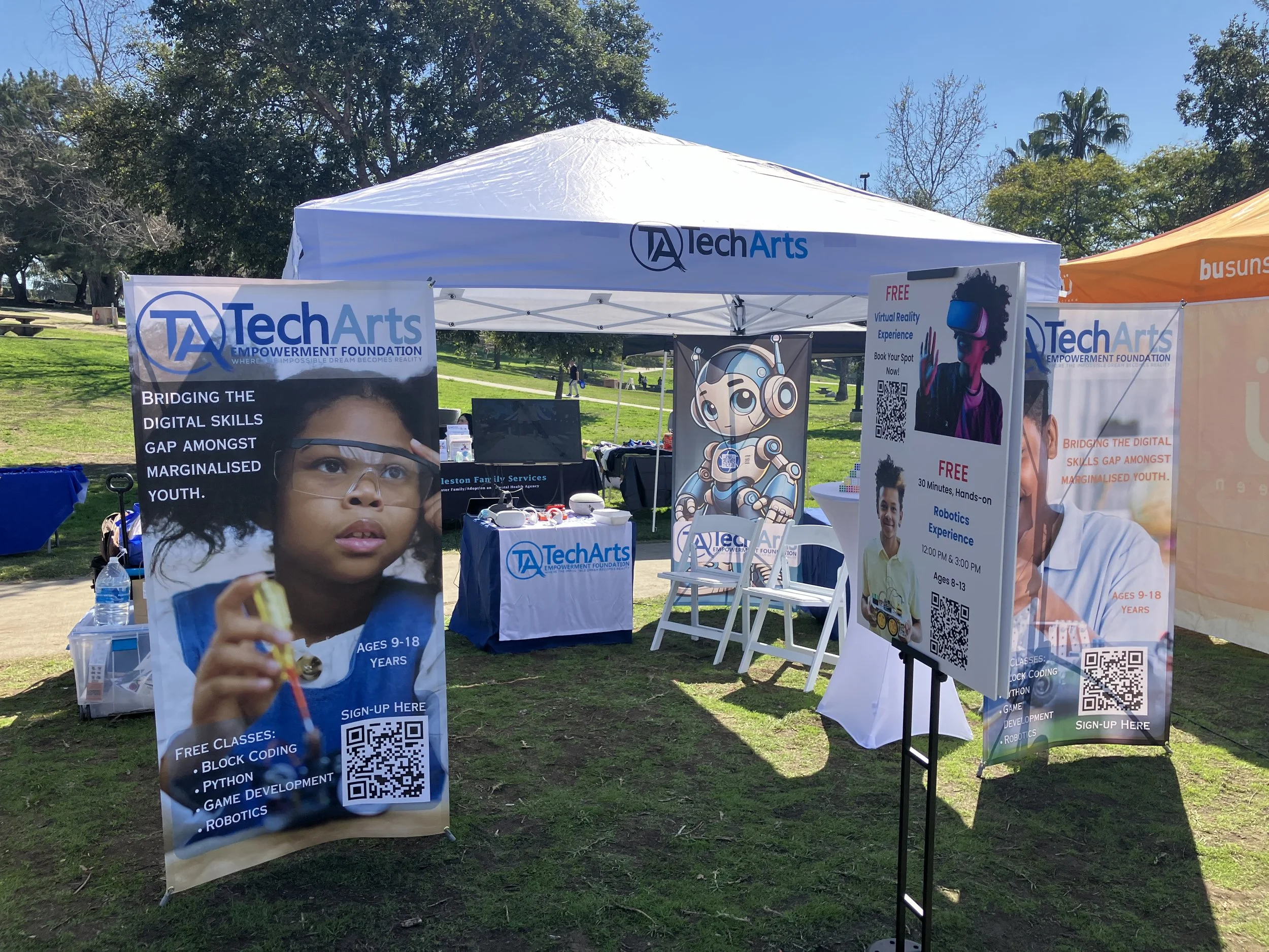 TechArts Empowerment Foundation booth at an outdoor event, featuring banners about digital skills for youth, virtual reality, and robotics experiences. The booth is set up with chairs and informational displays under a tent.