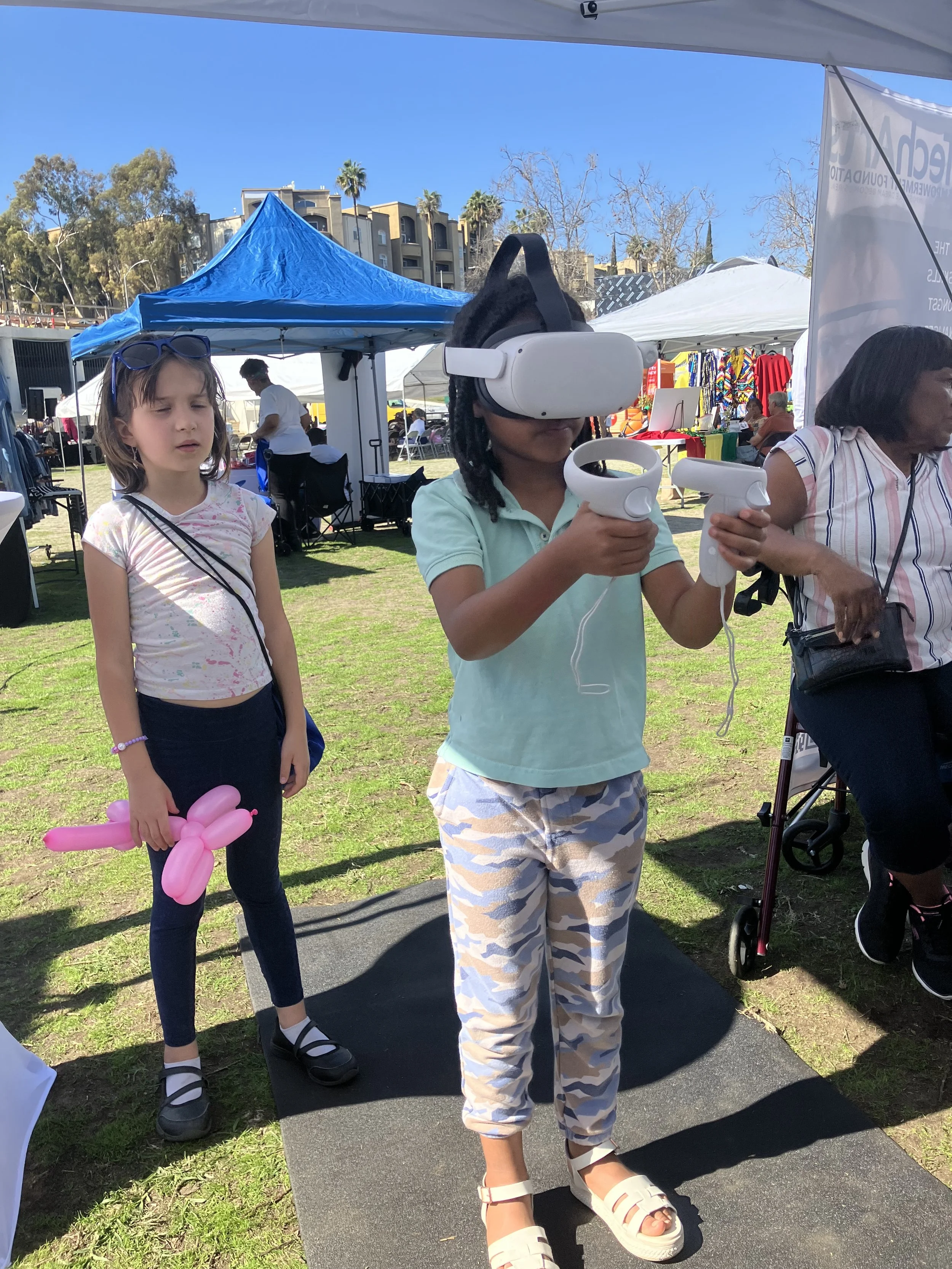 Two children participating in a virtual reality experience outdoors. One child wears a VR headset and holds controllers, while the other watches, holding a pink balloon animal. There are tents and people in the background, suggesting a festival or outdoor event.