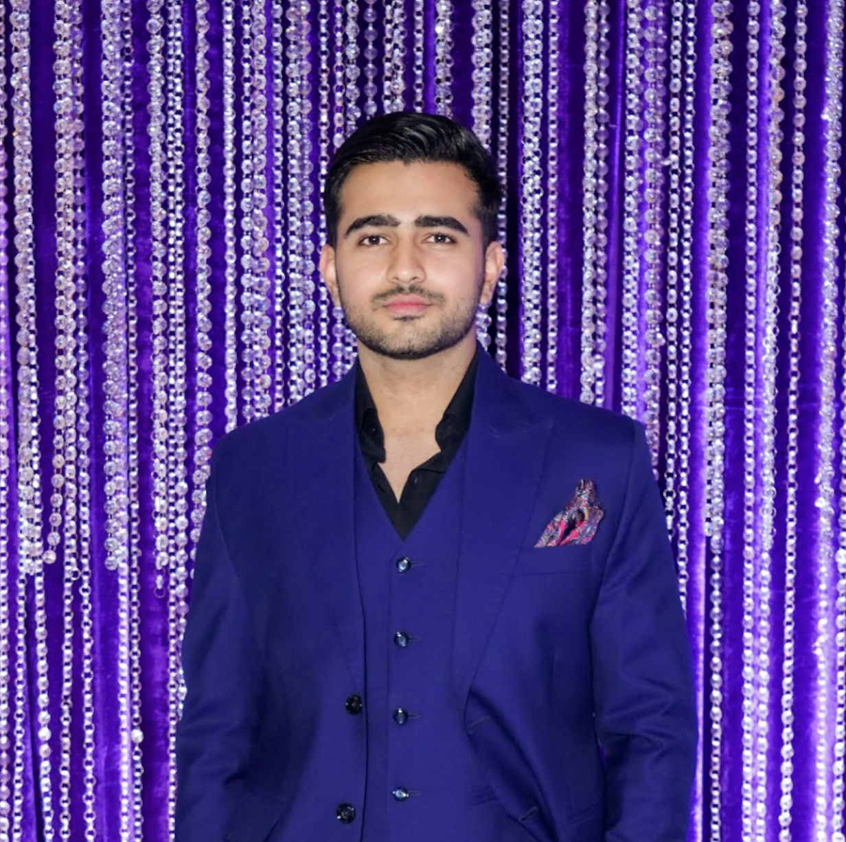 A young man in a navy blue suit with a pocket square, standing in front of a purple beaded curtain backdrop.
