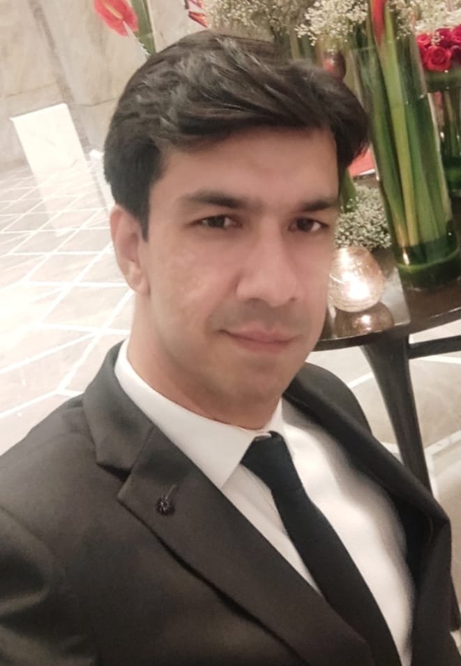 A man dressed in a black suit with a white shirt and black tie taking a selfie at a formal event or hotel, with floral arrangements in the background.