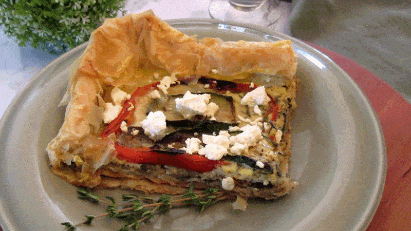 A slice of vegetable quiche with red peppers, eggplant, and feta cheese on a plate garnished with a sprig of thyme.