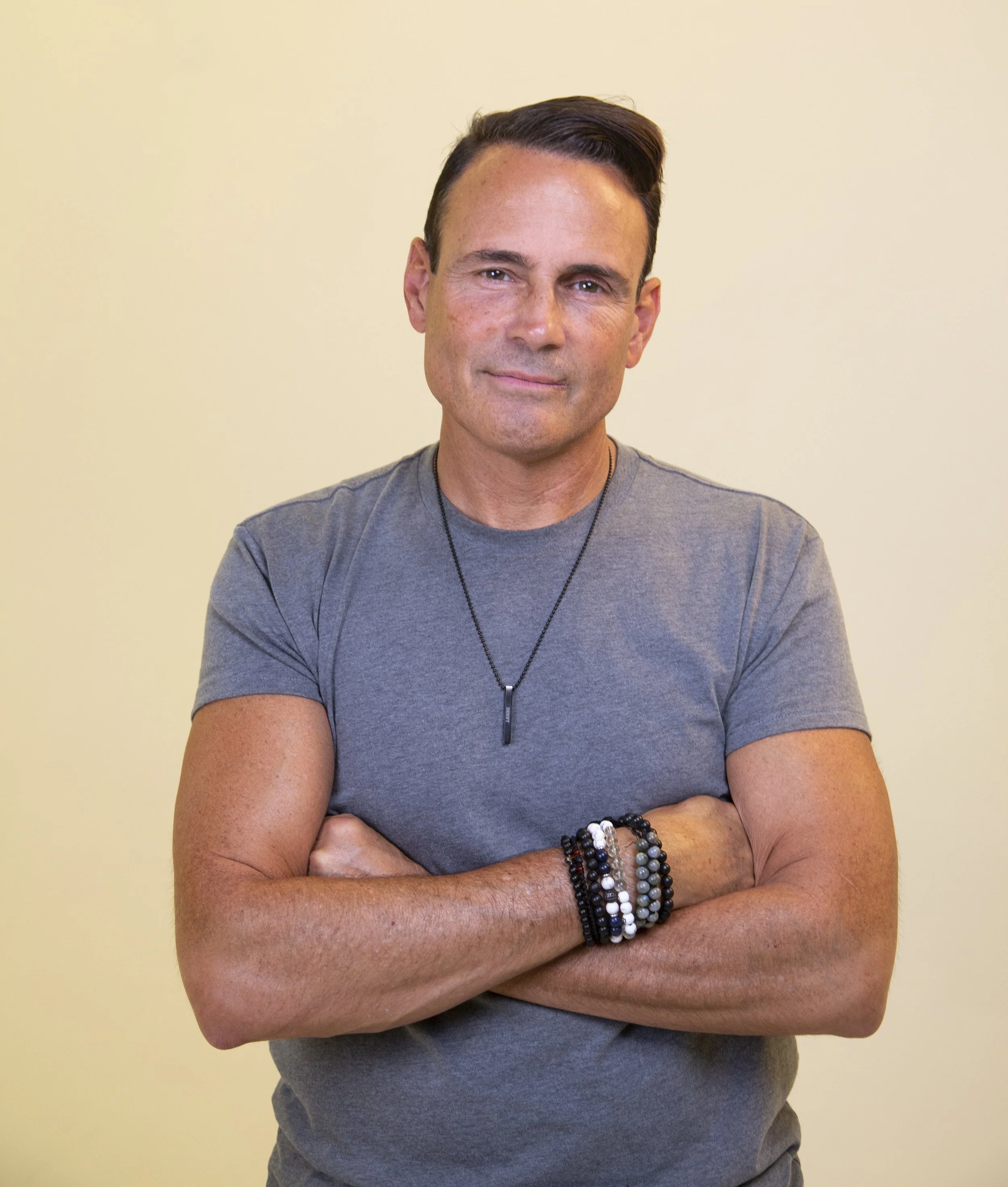 A man with short dark hair and light skin, wearing a gray t-shirt and multiple bracelets, standing with arms crossed against a plain yellow background.