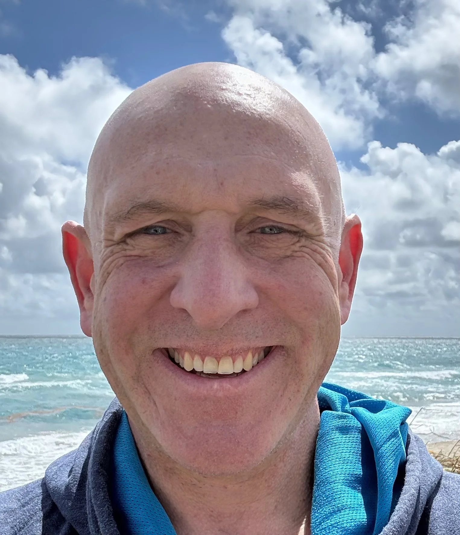 A smiling bald man taking a selfie at the beach, with the ocean and cloudy sky in the background.