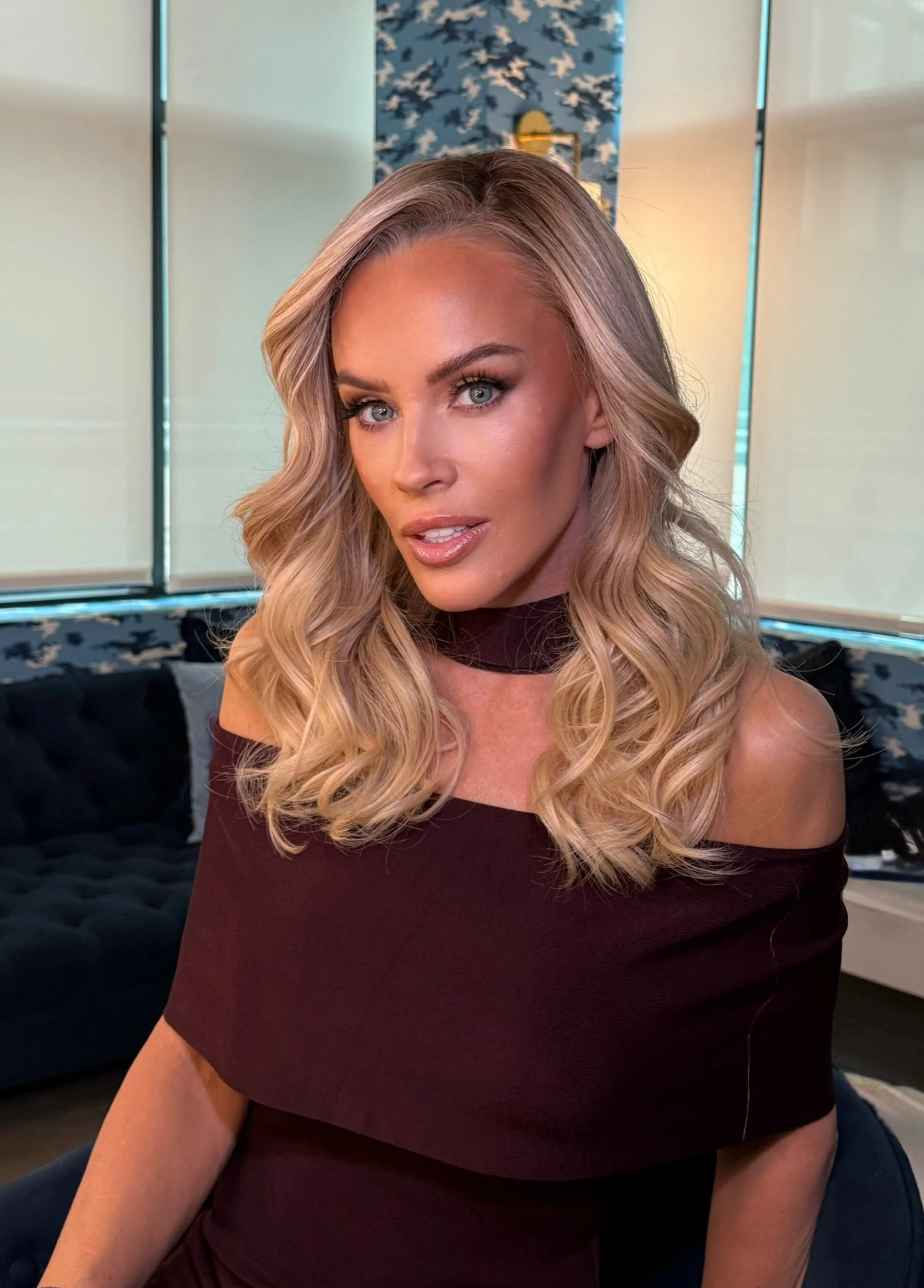 A woman with long, wavy blonde hair, blue eyes, and makeup, sitting indoors with a patterned blue and white wall behind her, wearing a black off-shoulder top.