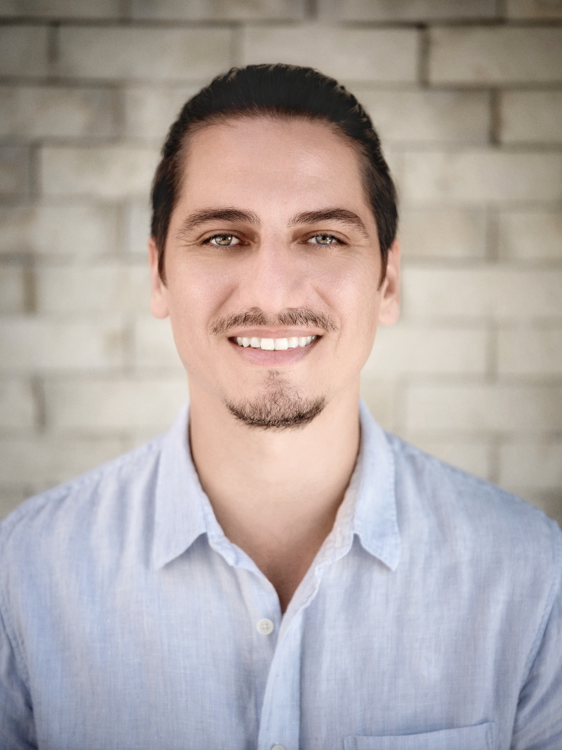 Portrait of a man with dark hair, light skin, a goatee, wearing a light blue button-up shirt, smiling, standing in front of a gray brick wall.