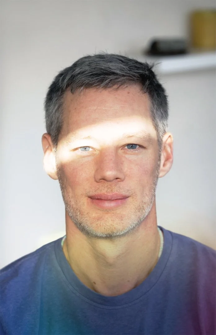 A man with short dark hair, blue eyes, and light facial hair is shown in a close-up portrait. Sunlight partially illuminates his face. He is wearing a dark blue shirt, and the background includes a blurred shelf with a yellow container and other objects.
