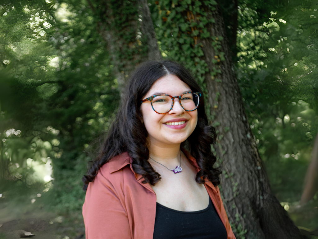 A smiling young woman with dark curly hair wearing glasses, a black top, and a rust-colored jacket, standing outdoors in front of a tree with green foliage.