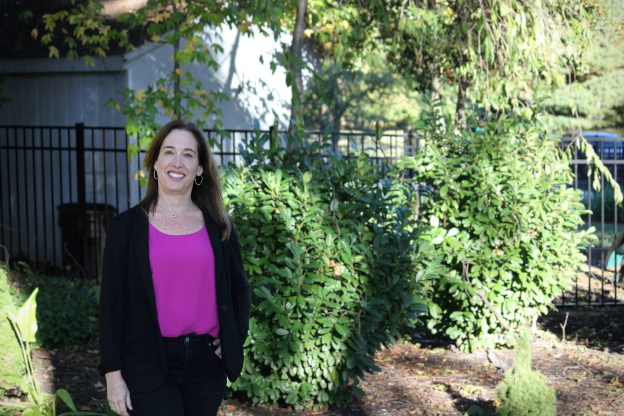 A woman with brown hair, wearing a black blazer over a bright pink top, standing outdoors in front of lush green bushes and trees, smiling at the camera.