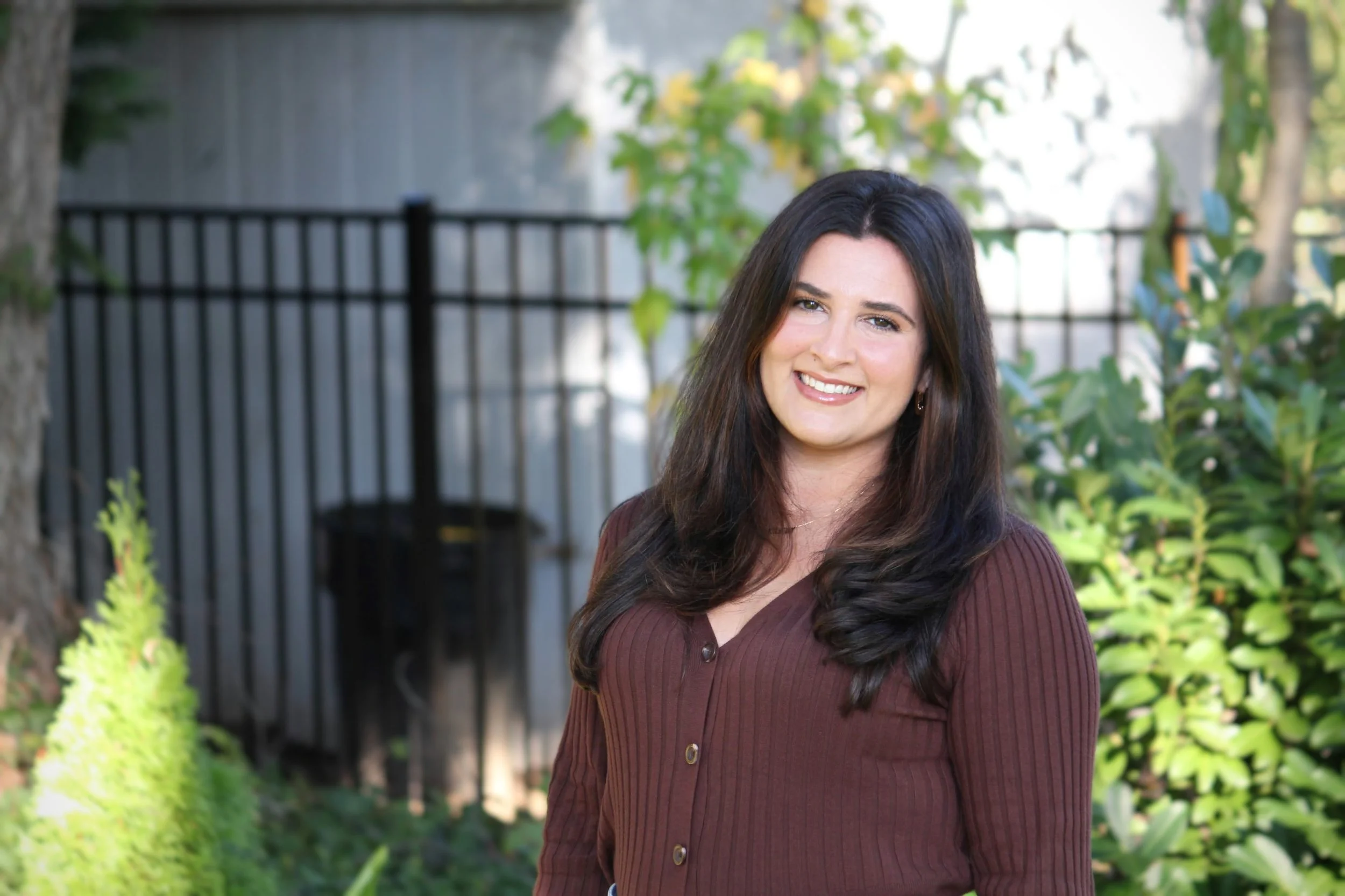 A woman with long, dark brown hair smiling outdoors, wearing a brown button-up shirt, with greenery and a black metal fence behind her.