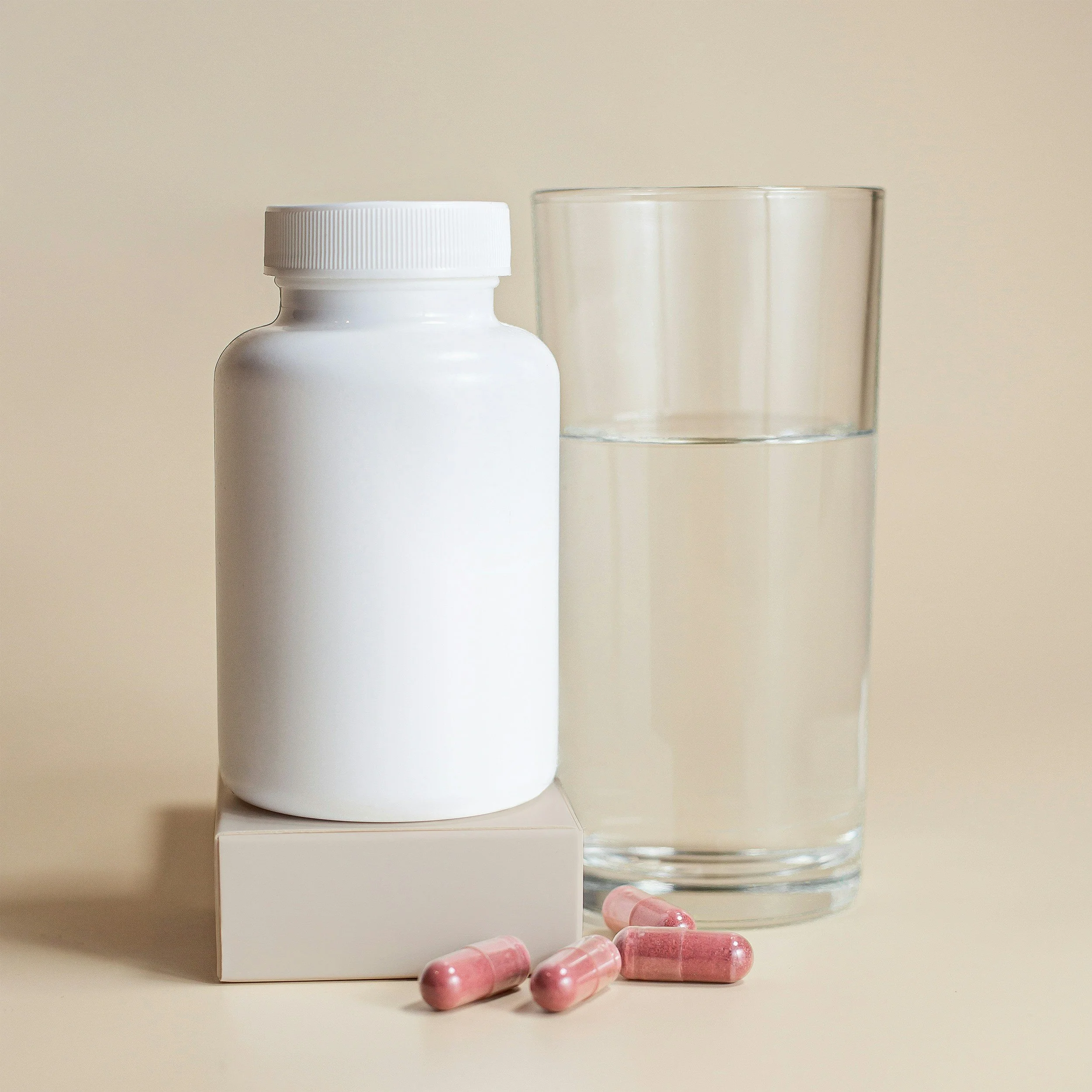 A white medication bottle, a glass of water, and pink capsules on a neutral background.