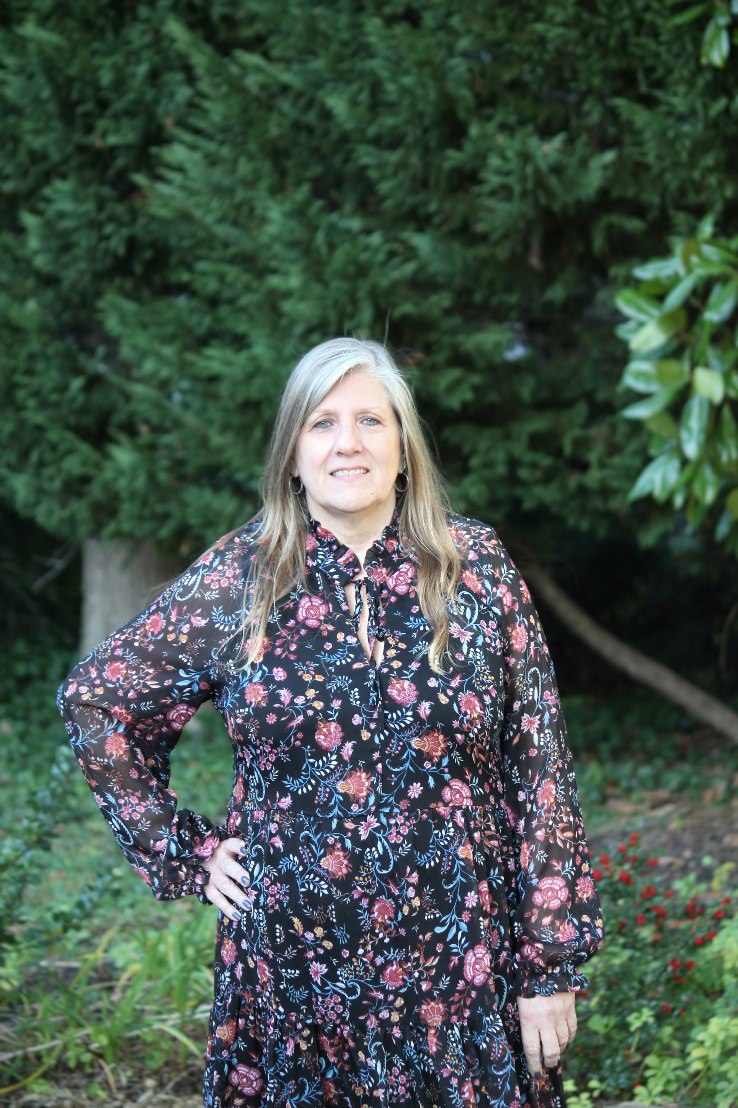 A woman standing outdoors in front of green trees and bushes, wearing a black floral dress with long sleeves.