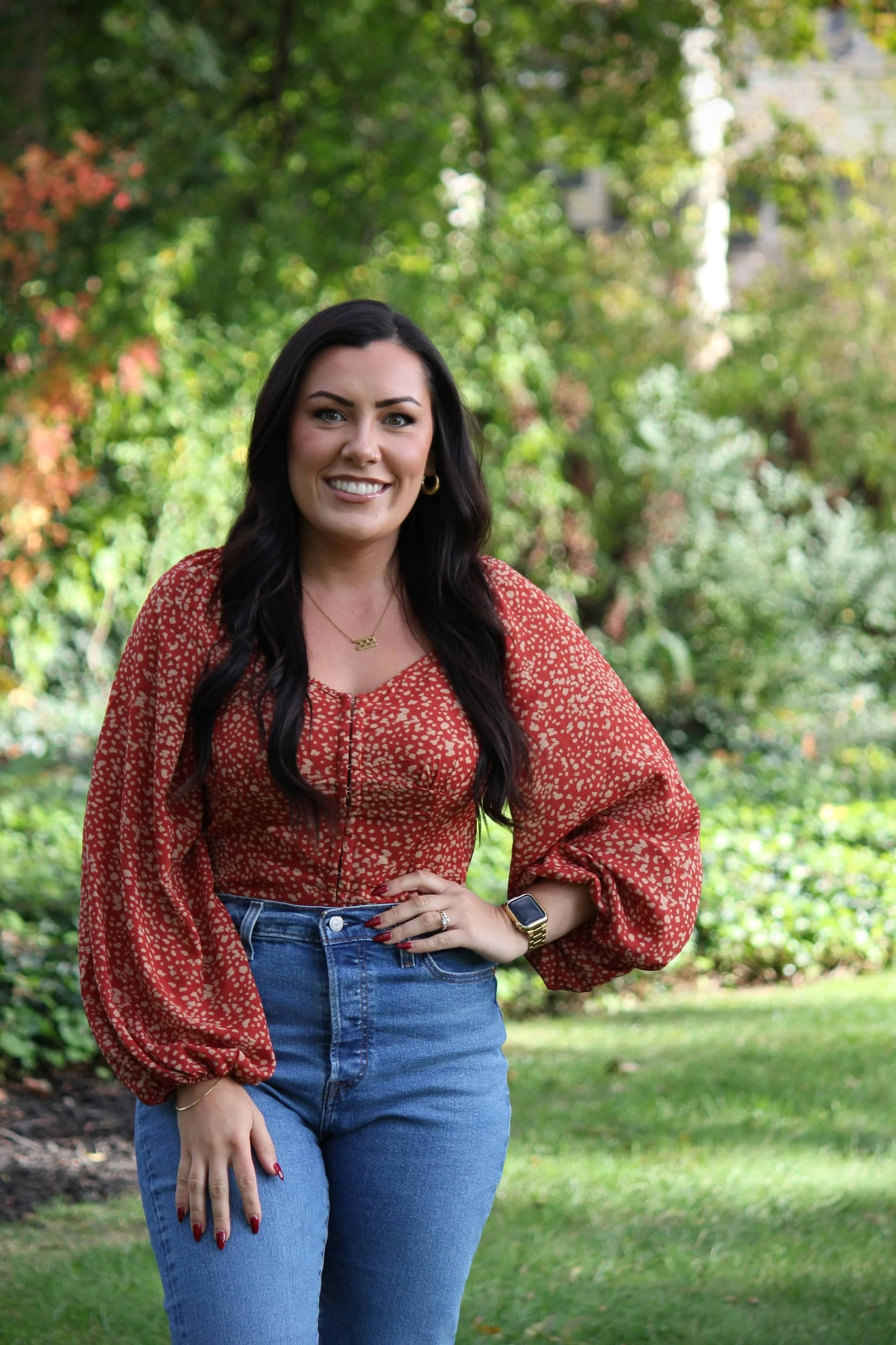 A woman standing outdoors in a garden with lush green trees and plants, smiling and posing for the camera.