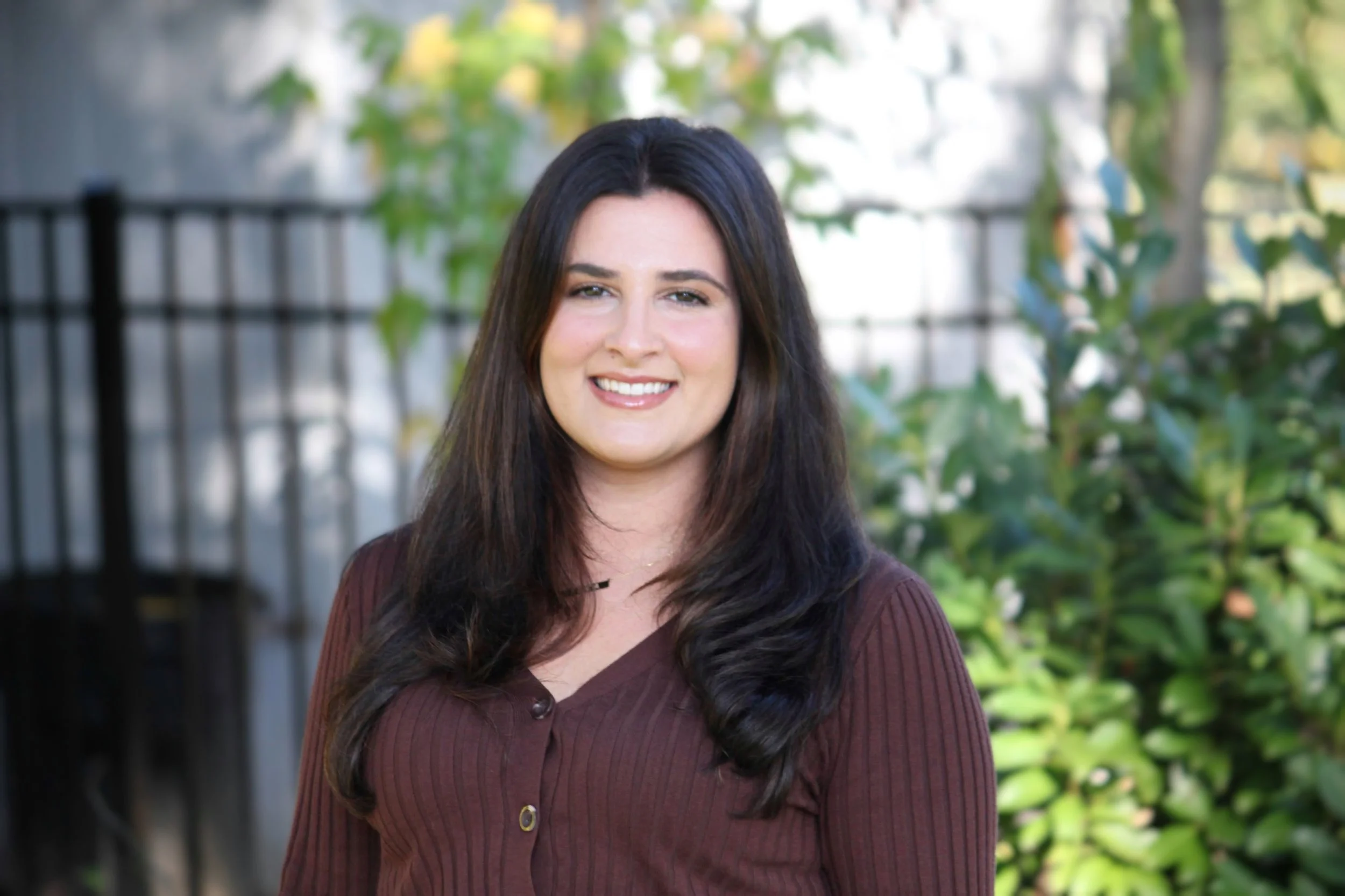 A woman with long dark hair wearing a maroon shirt smiling outdoors with a background of green trees and a black metal fence.