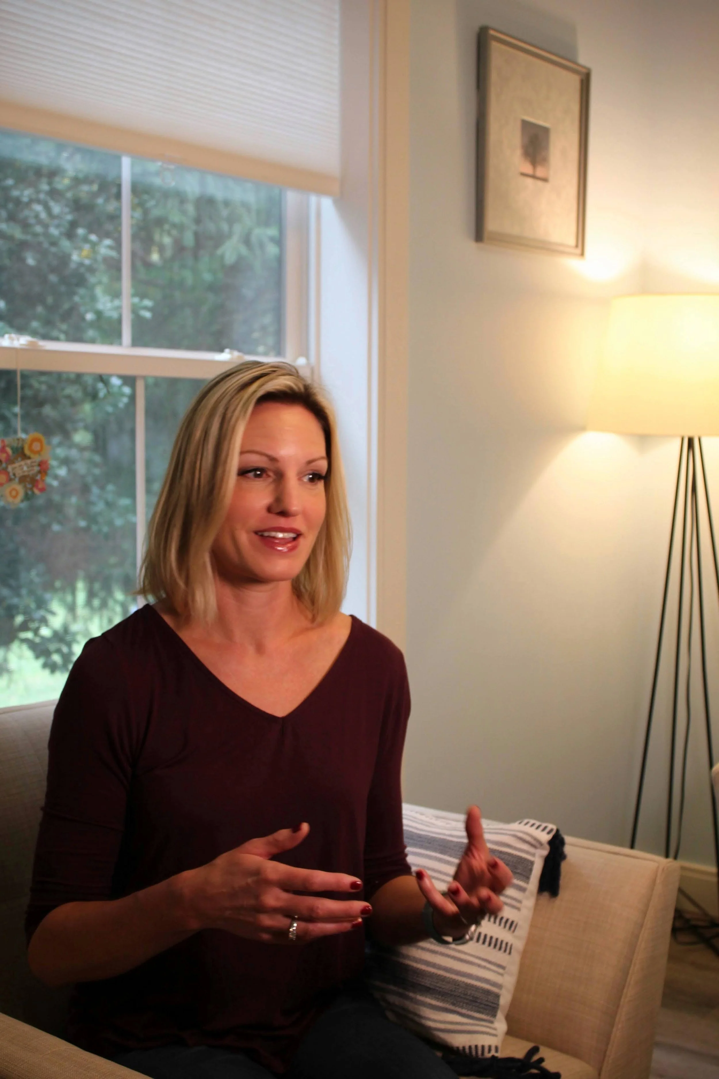 A woman with blonde hair wearing a dark red top is sitting on a beige couch, talking with her hands. There is a window with a porch and greenery outside, a framed picture on the wall, and a floor lamp in the background in a cozy living room.