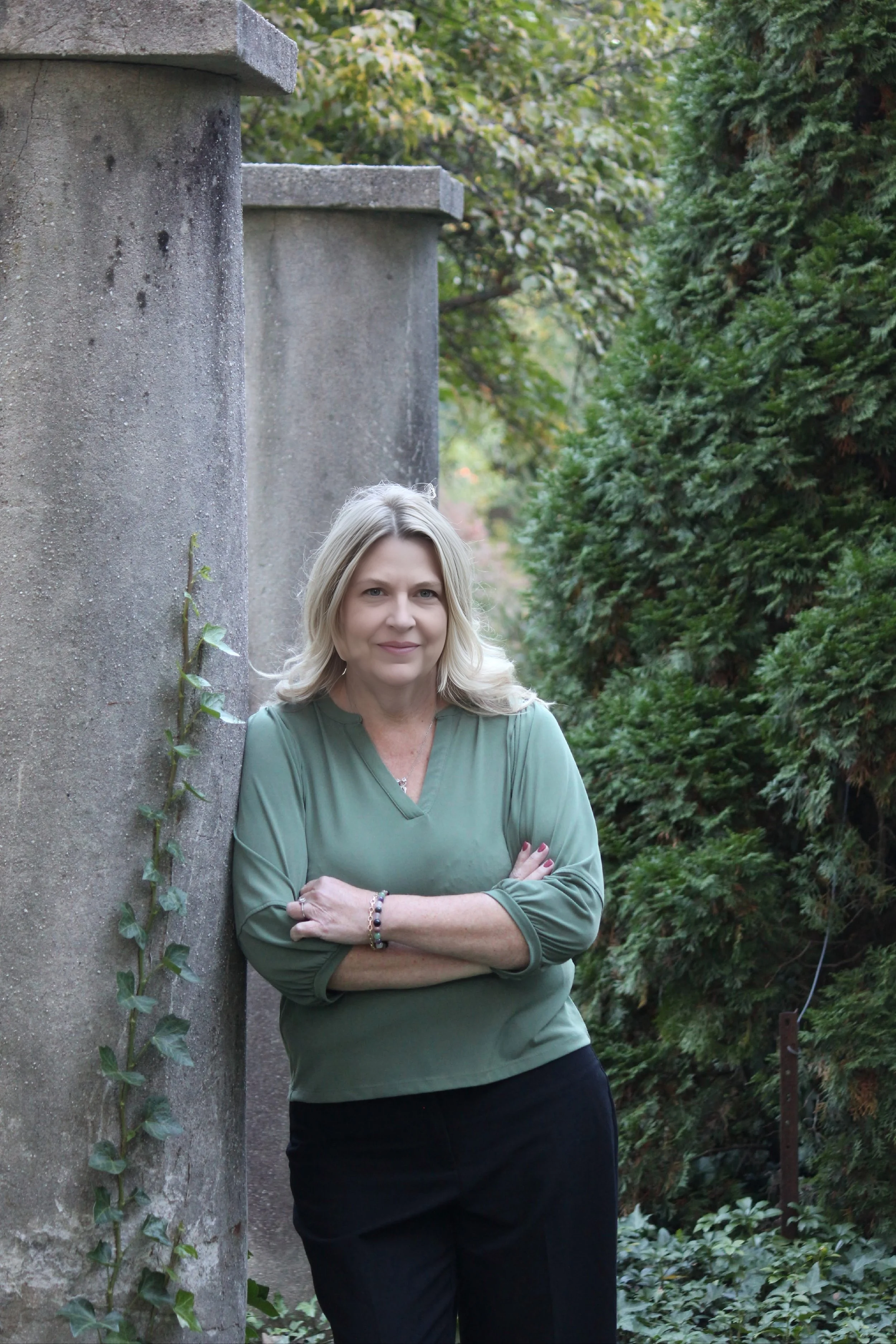 A middle-aged woman with blonde hair, wearing a green blouse and black pants, standing outdoors with her arms crossed, leaning against a concrete wall, surrounded by green foliage.