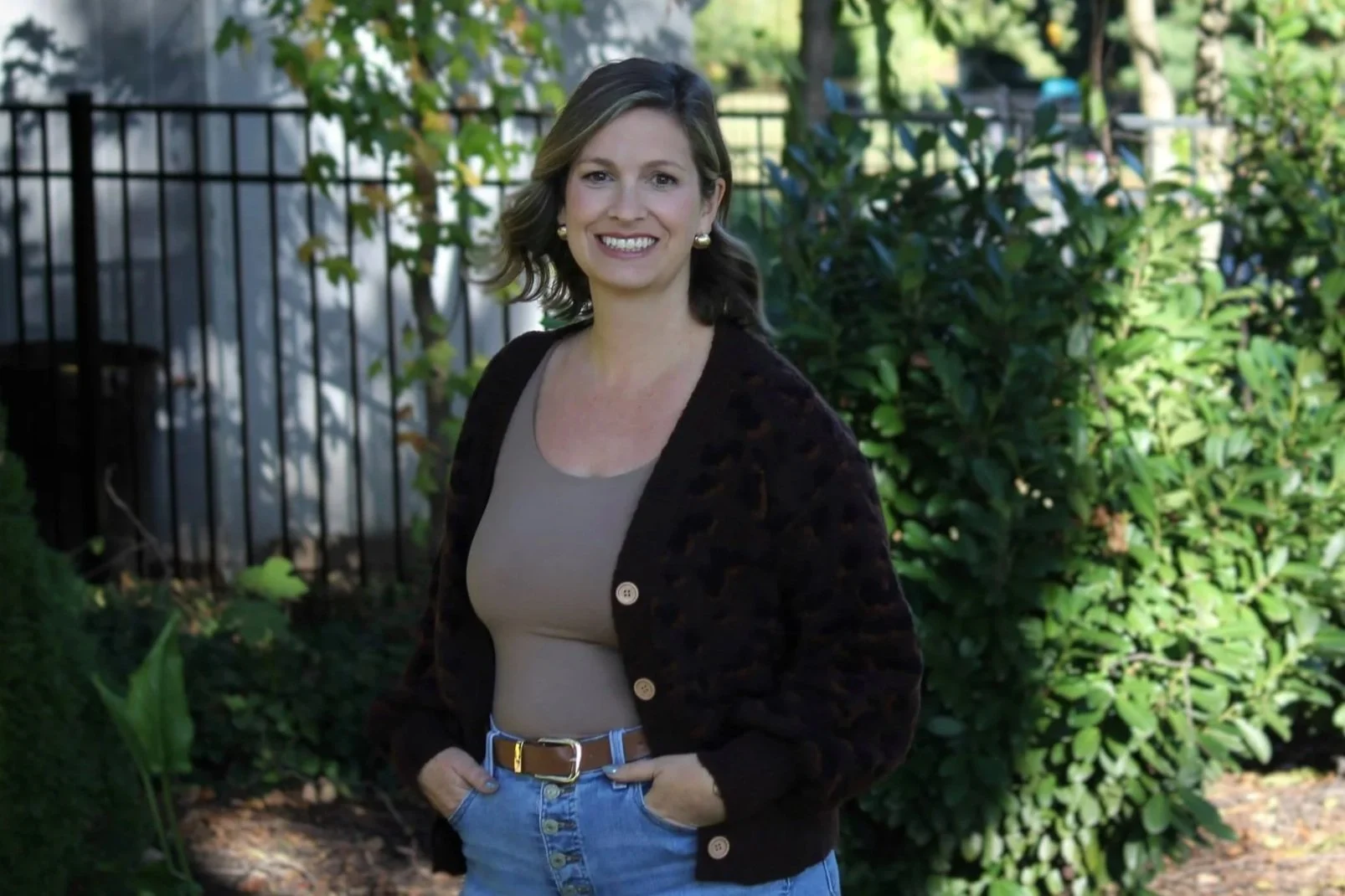 A woman with shoulder-length brown hair smiling outdoors, wearing a beige top, a black cardigan, and blue jeans, standing in front of green bushes and a black metal fence.