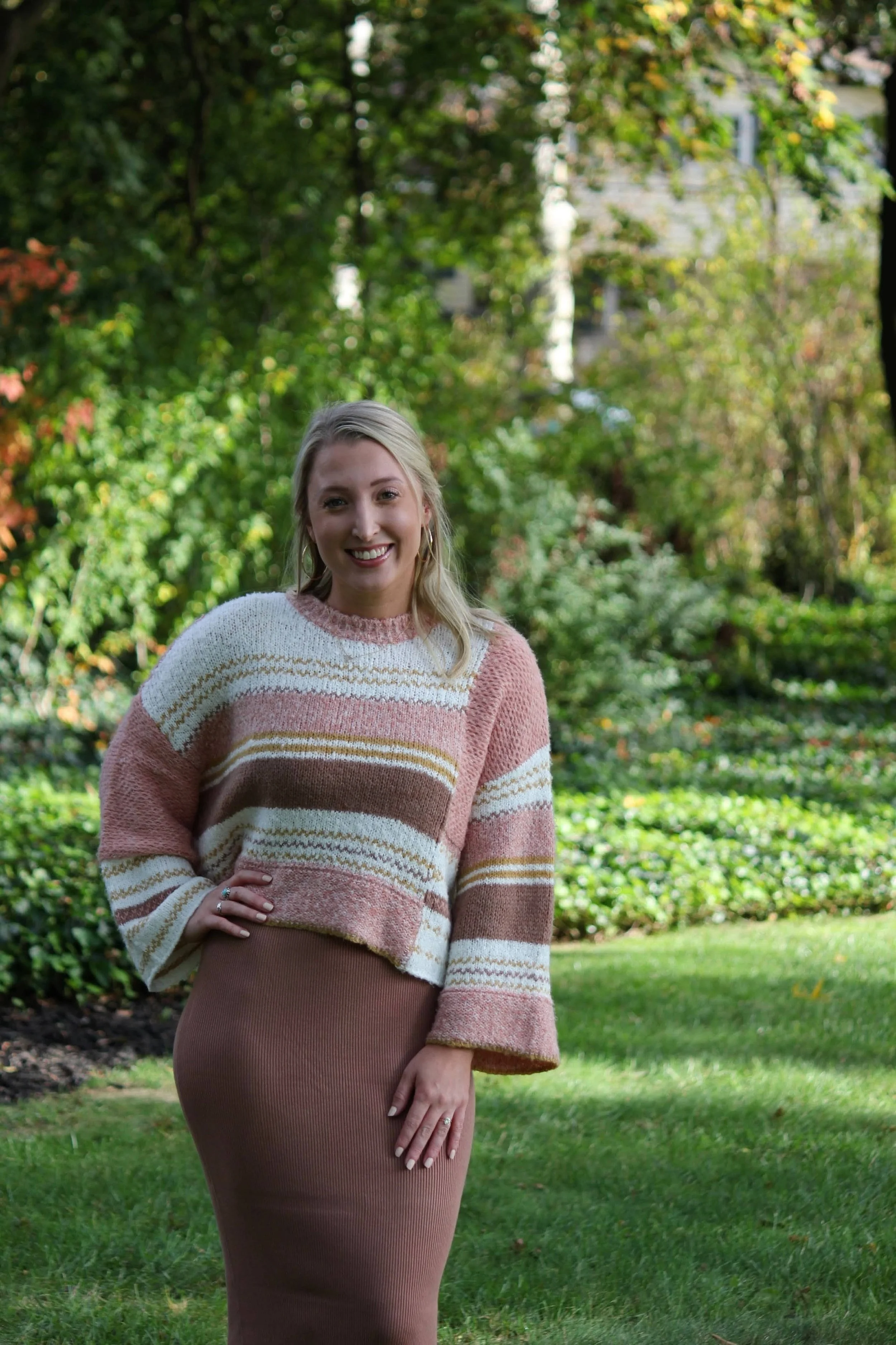 A young woman with blonde hair wearing a pink, cream, brown, and yellow striped knit sweater and a brown ribbed skirt, standing outdoors in a lush garden with greenery and trees in the background.