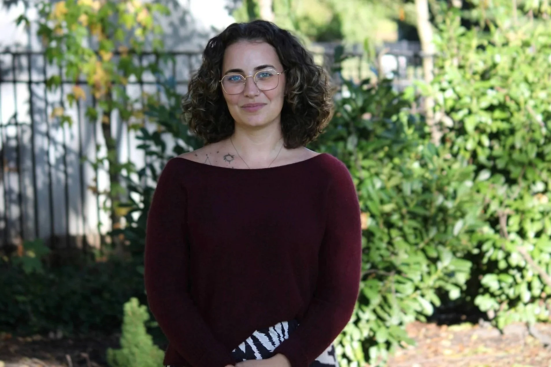 A woman with curly dark hair, glasses, and a tattoo on her shoulder stands outdoors in front of green bushes, wearing a maroon long-sleeve shirt.