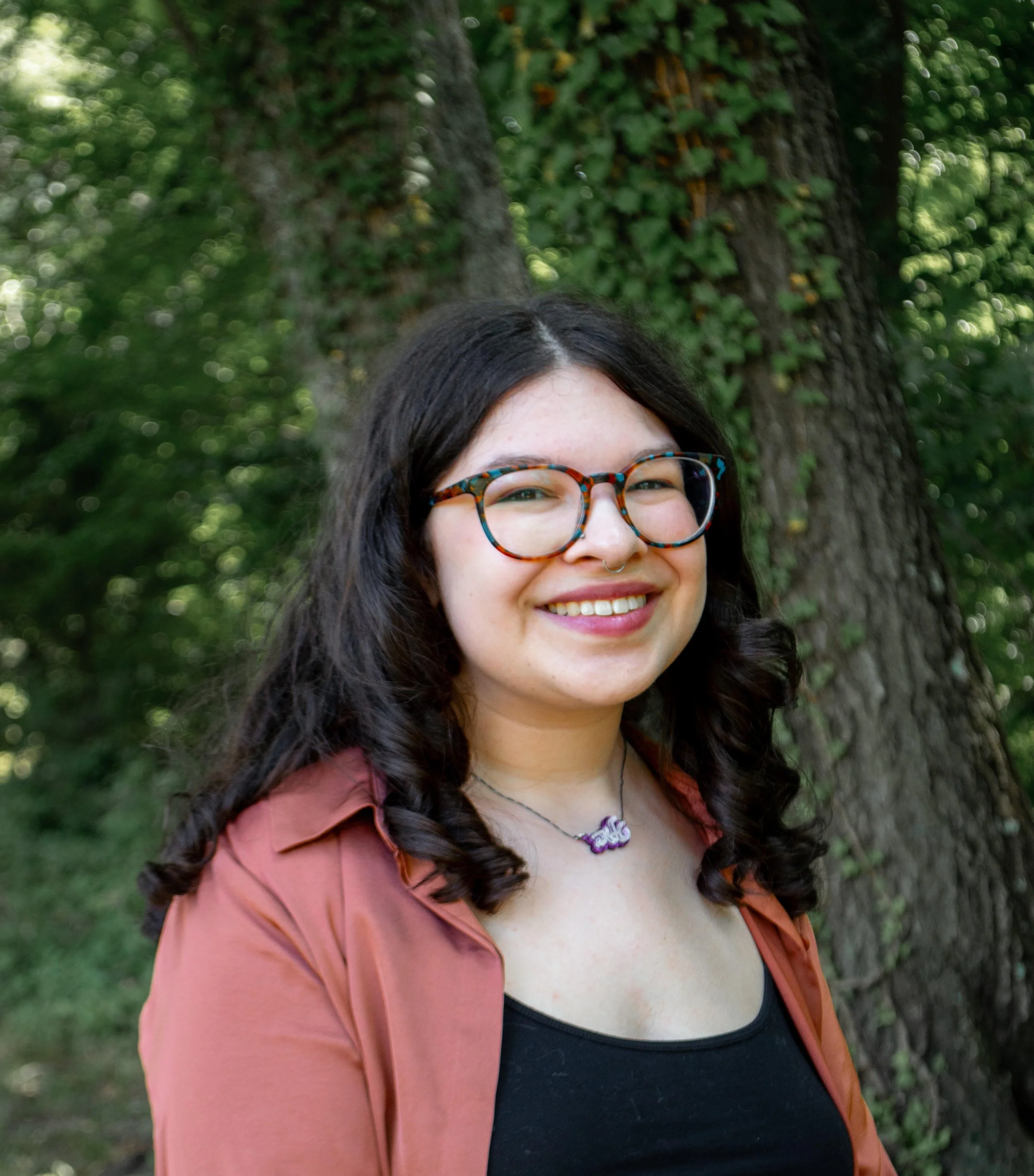 Young woman with dark curly hair, colorful glasses, and a septum piercing smiling outdoors in front of a large tree with green foliage.