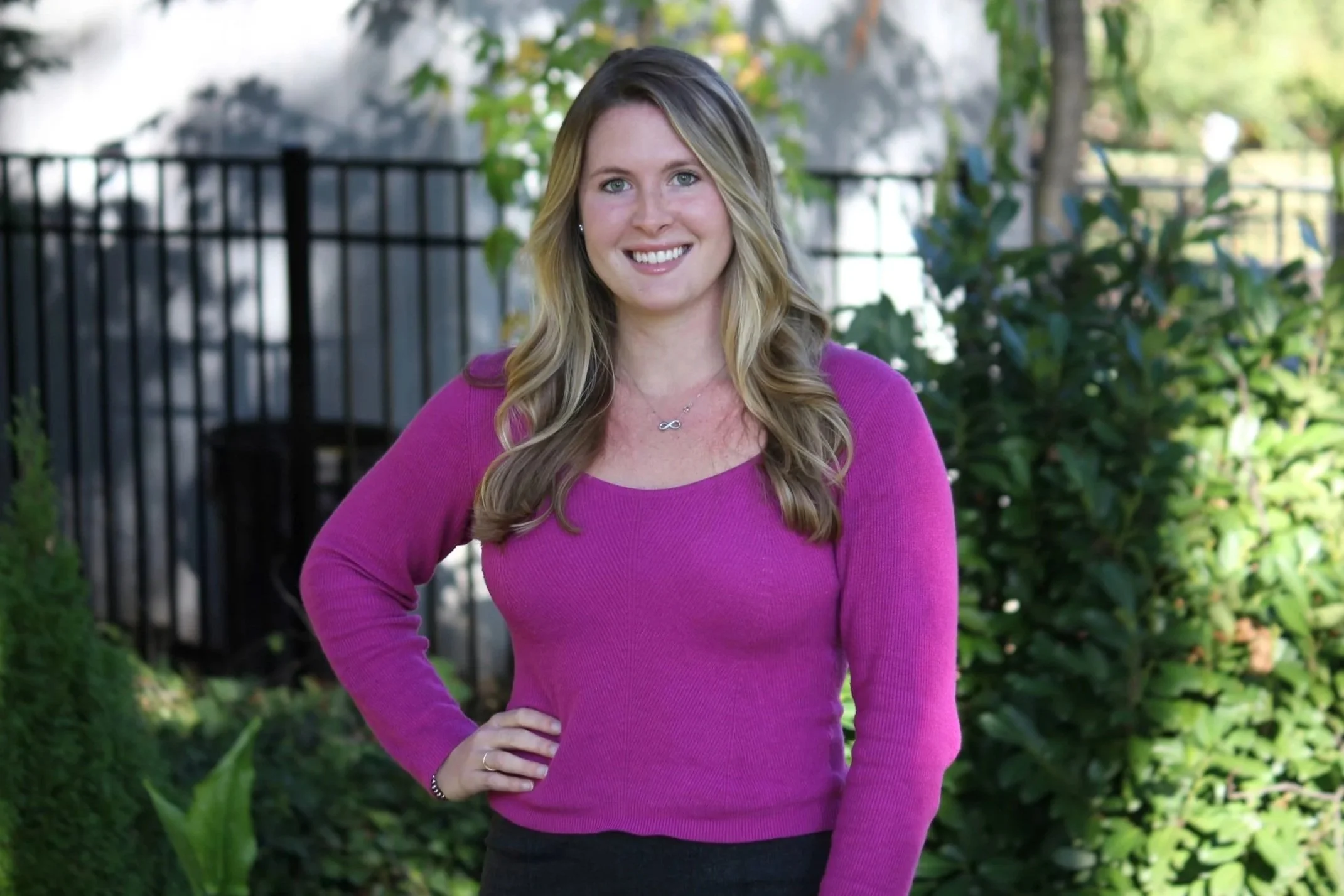 A woman with long blond hair wearing a bright pink sweater, standing outdoors with green plants and a metal fence in the background, smiling at the camera.