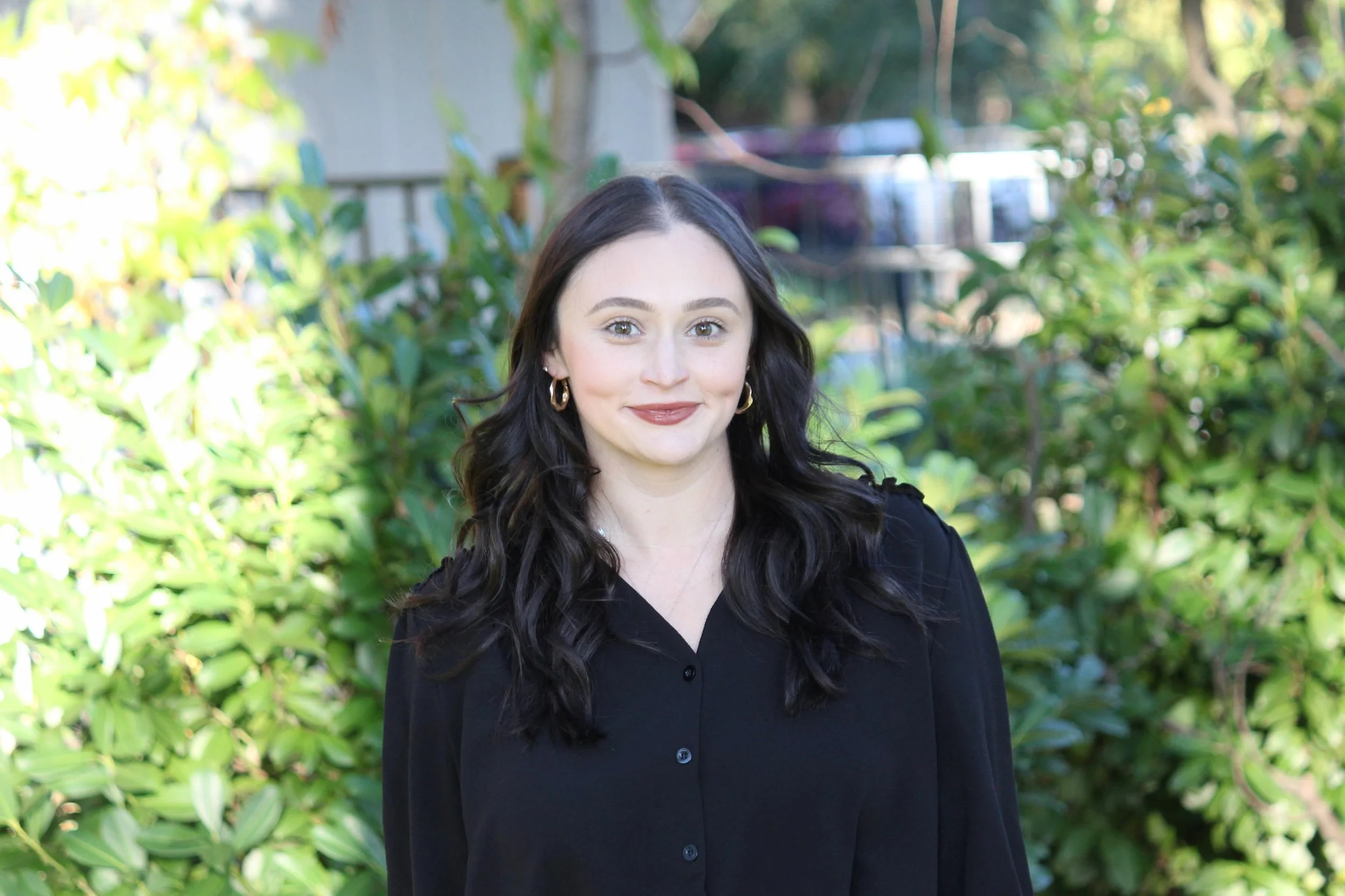 A young woman with long, wavy dark hair, wearing a black button-up shirt, standing outdoors in front of lush green foliage, smiling at the camera.