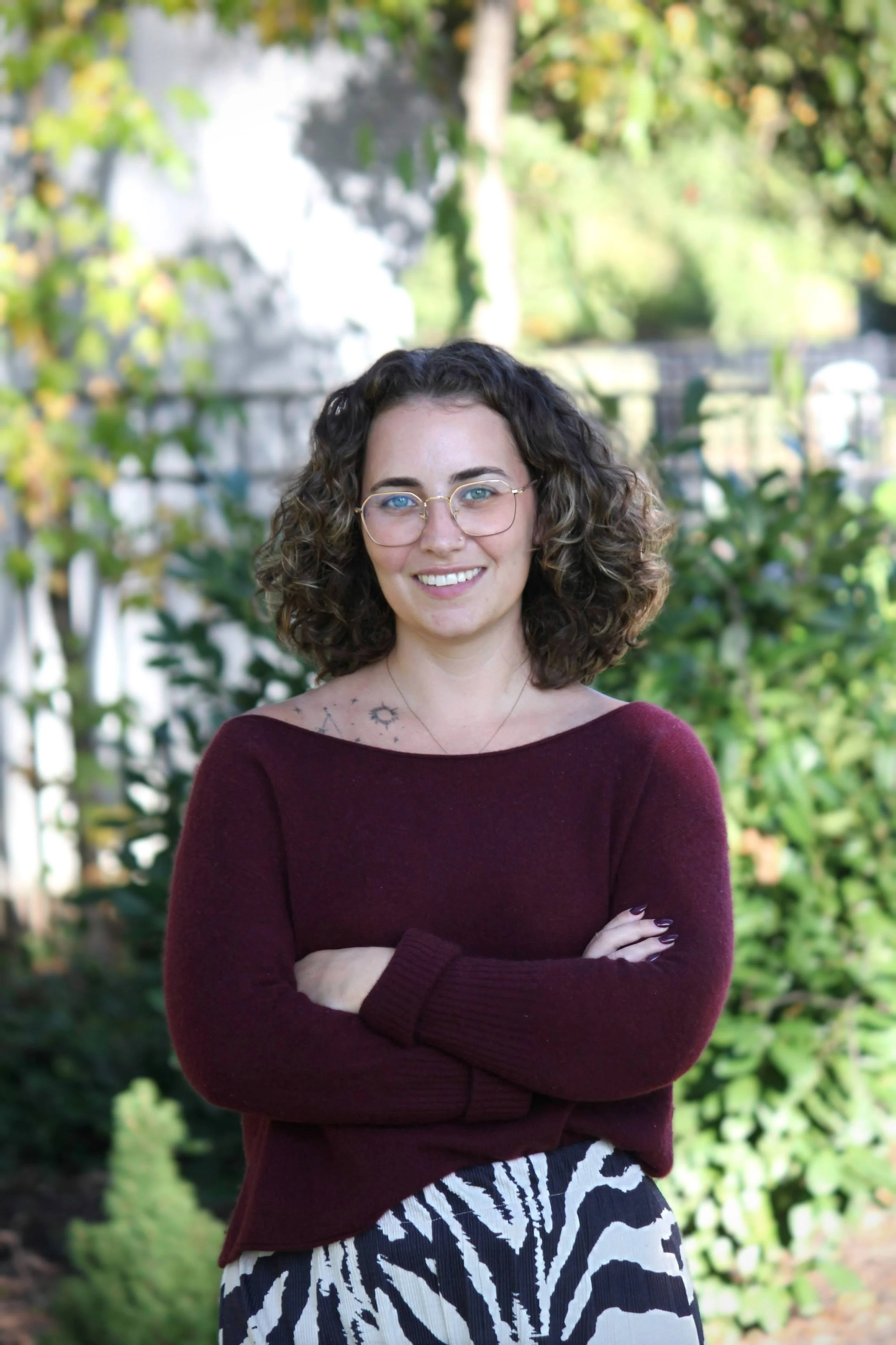 A woman with curly brown hair, glasses, and a tattoo on her chest, standing outdoors with arms crossed, wearing a maroon sweater and zebra print pants, smiling with a background of trees and greenery.