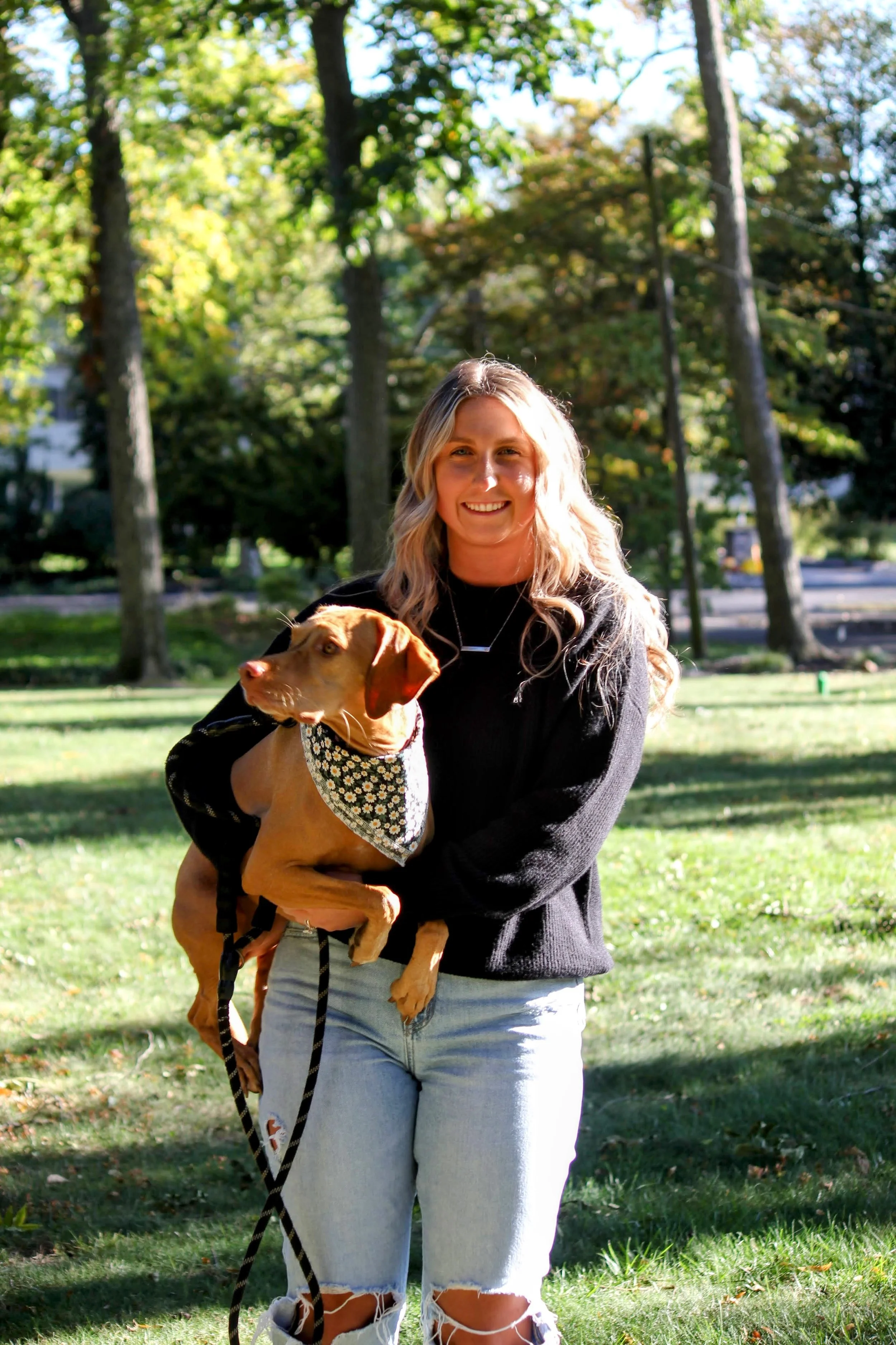 Woman holding a dog outdoors in a park with trees and grass, smiling at the camera.