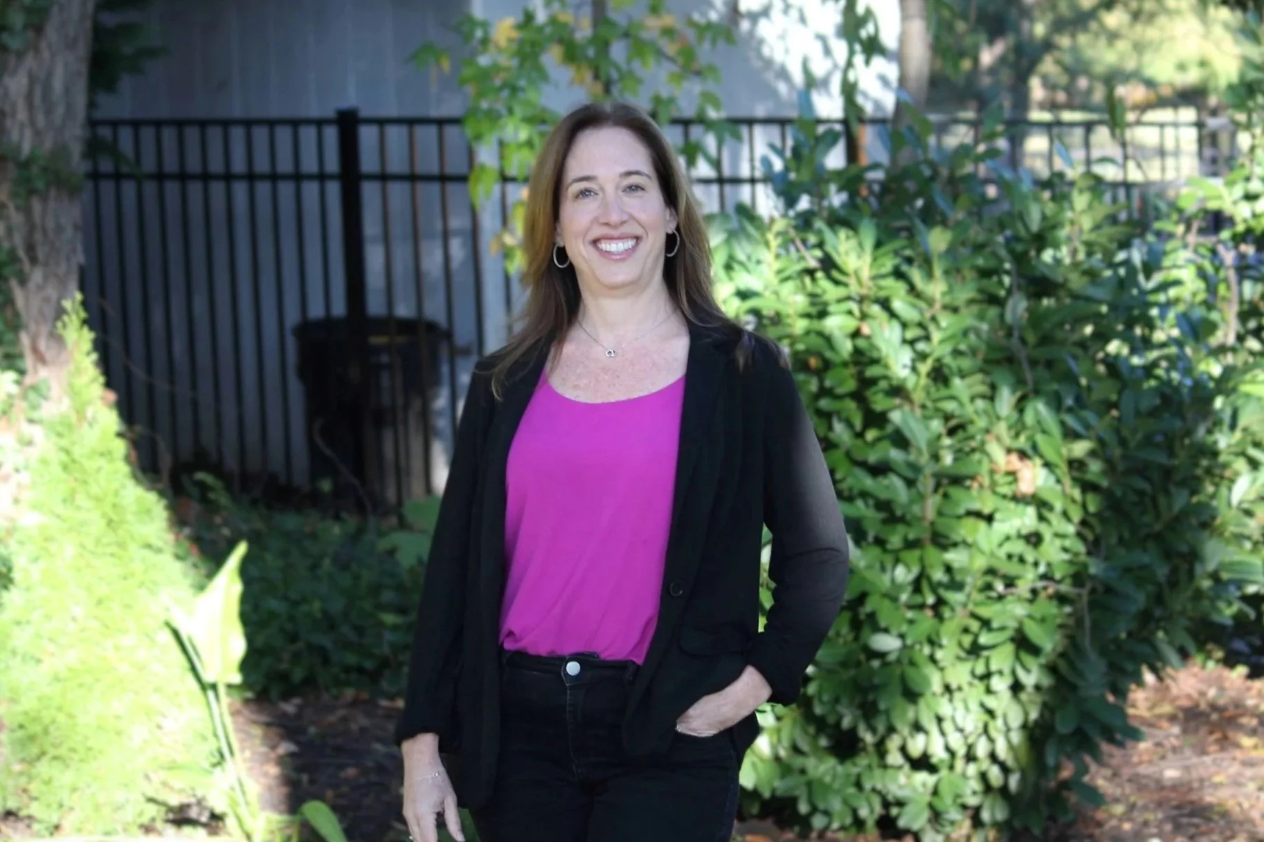 A woman with brown hair in a black blazer and purple top, smiling outdoors in front of green bushes and trees.