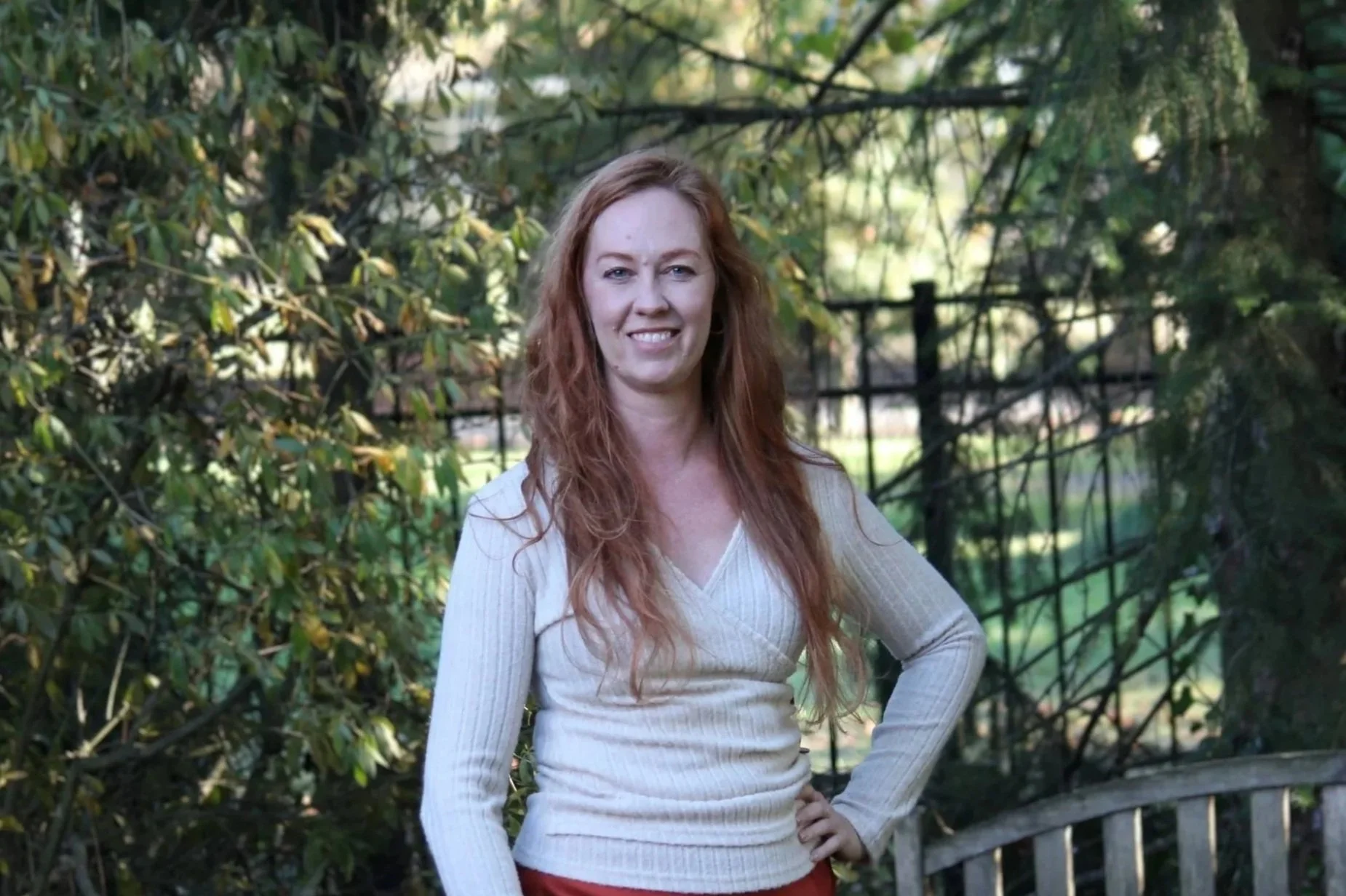 A woman with wavy brown hair in a professional outfit standing outdoors with a background of green trees and bushes.