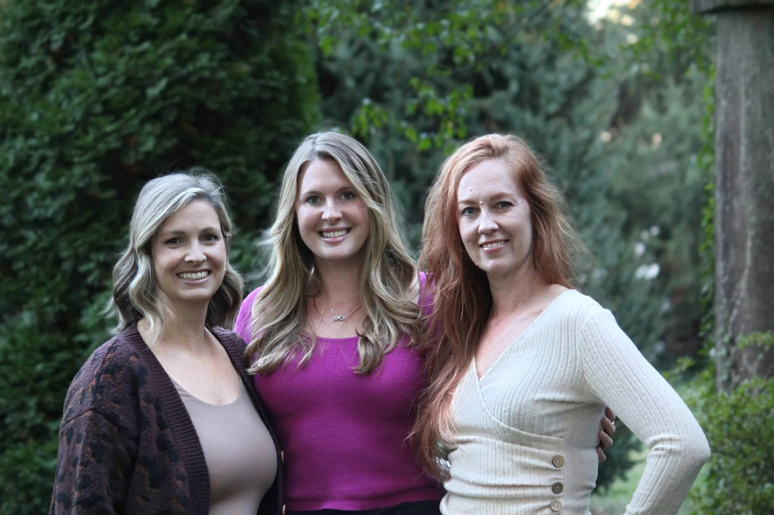 Three women smiling outdoors in front of a green, leafy background.