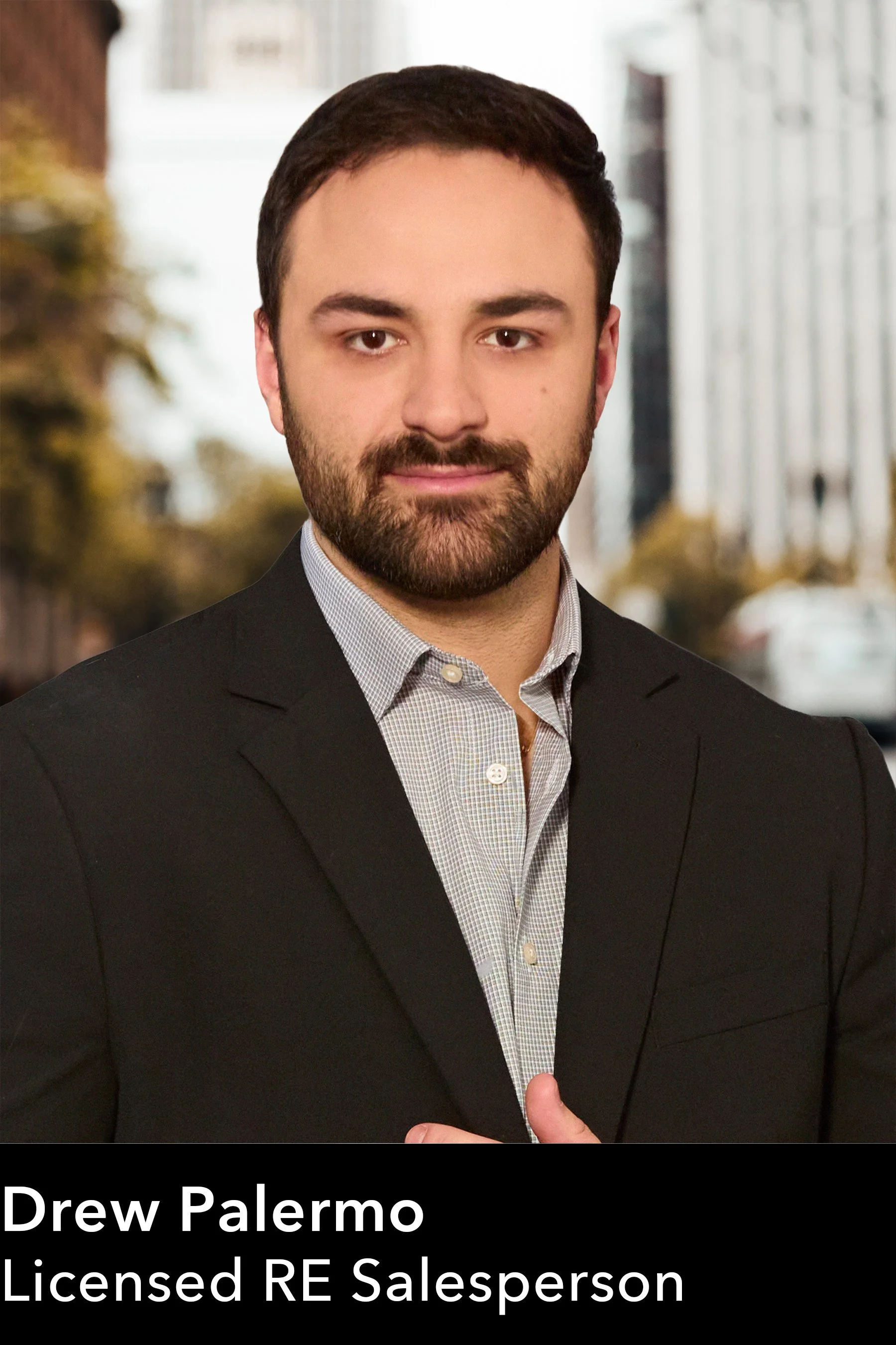 A man in a black blazer and light-colored shirt standing outdoors in front of a blurred background of buildings and trees.
