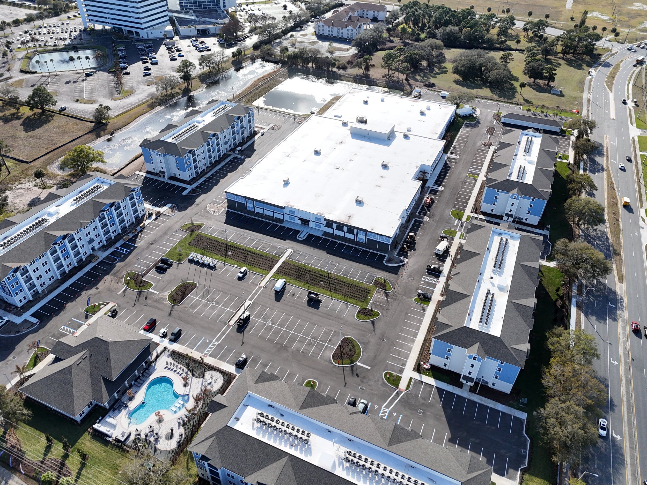 Aerial view of a suburban area with multiple buildings, parking lots, a swimming pool, small lakes, trees, roads, and open grassy fields.
