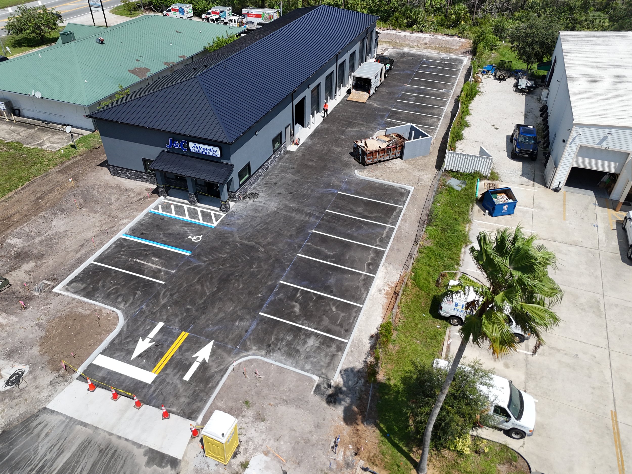 Aerial view of a parking lot with designated parking spaces, some under construction, outside a building labeled 'J&C Automotive Repair & Service'. There are construction cones and equipment, and a person in a safety vest is near the building's entrance.