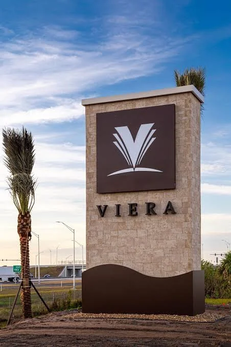 A tall stone monument with a dark rectangular sign displaying a stylized open book logo and the word 'VIERA' below. Two palm trees and a blue sky with clouds are in the background.