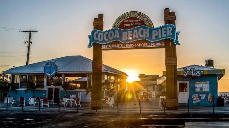 Cocoa Beach Pier entrance sign at sunset with restaurants, bars, shops, and fishing, and a seafood restaurant named Sea Dog's.