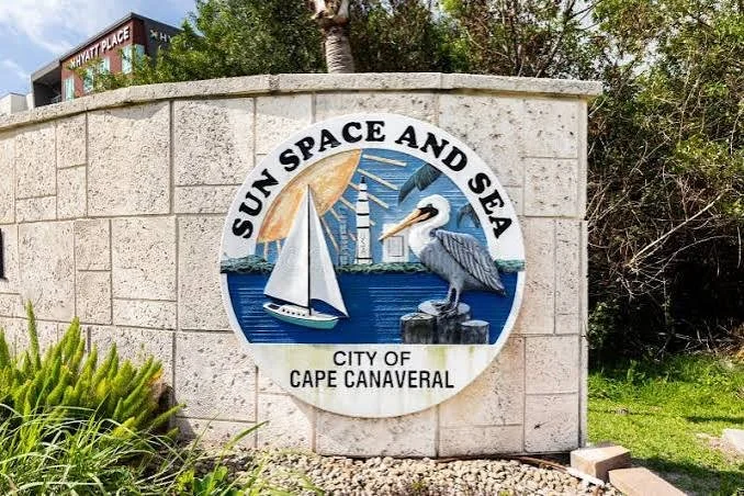A circular emblem on a stone wall with the text "Sun Space and Sea" and "City of Cape Canaveral." The emblem features a sailboat, a lighthouse, and a pelican against a backdrop of the sun and ocean.