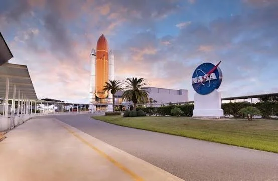 NASA logo on a standee and a space shuttle replica outdoors, with a cloudy sky at sunset.