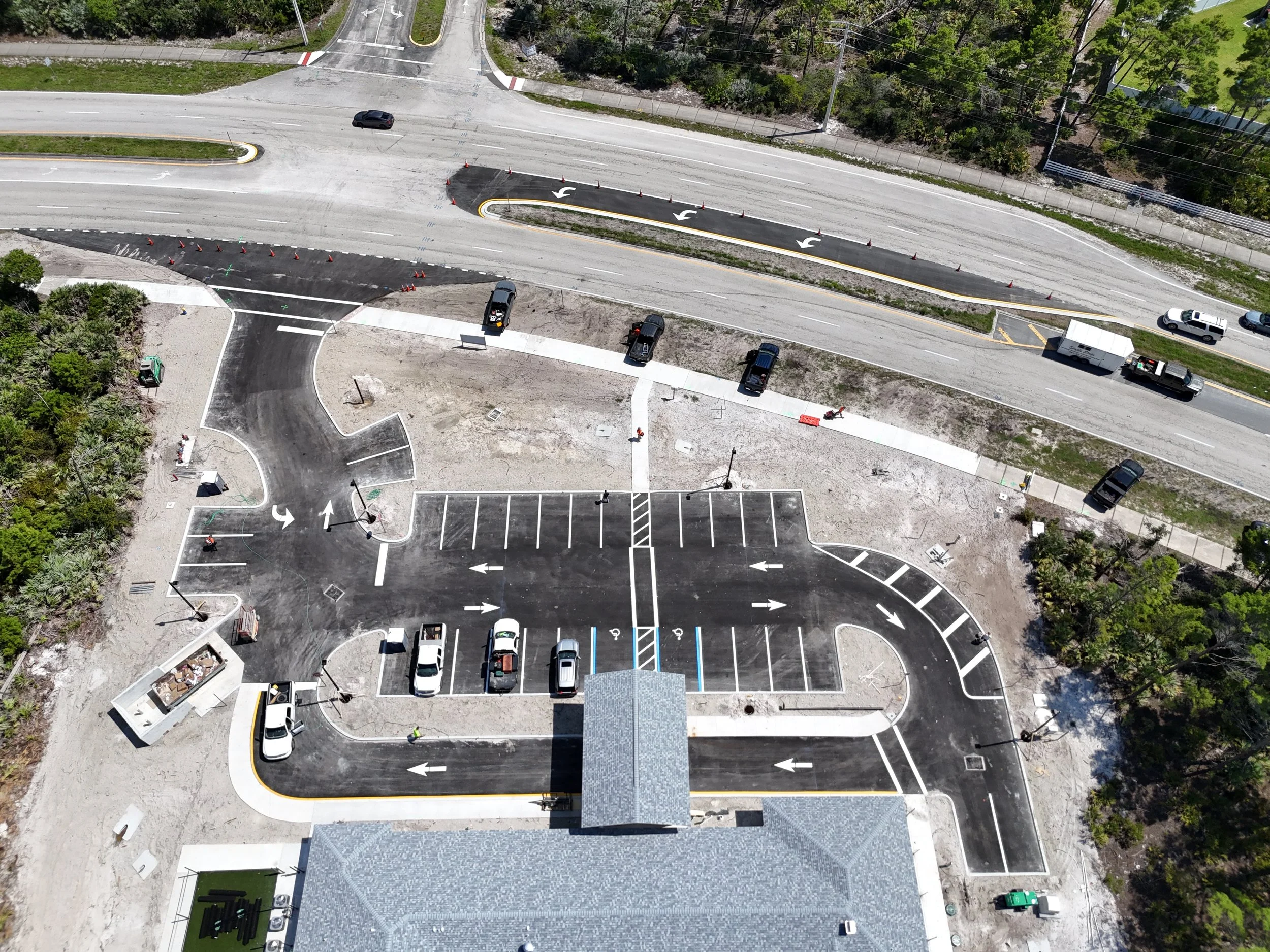 An aerial view of a parking lot and road under construction, with parked cars, construction cones, and freshly paved sections.